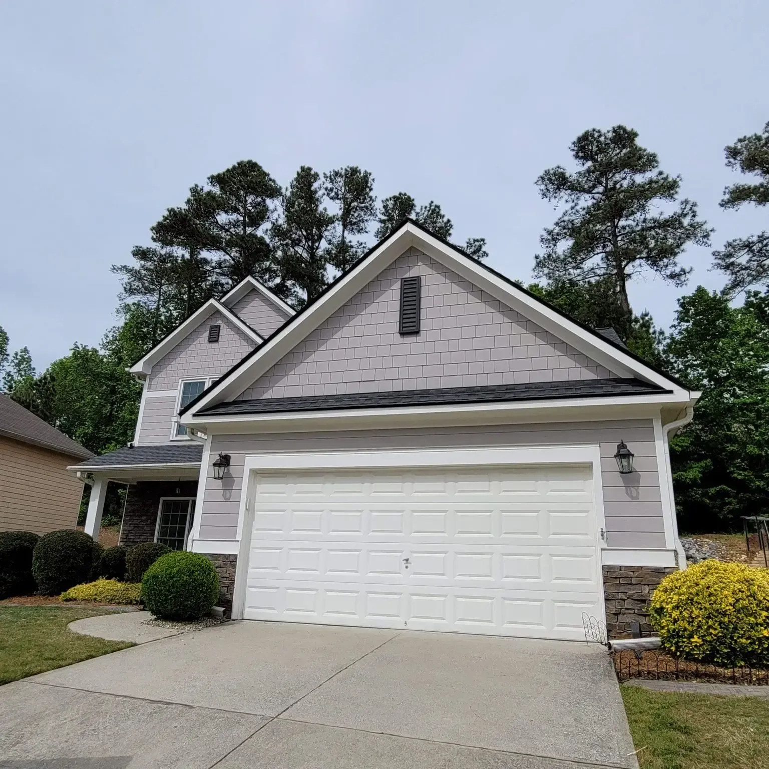 Two-story house with gray siding, white garage door, and black roof, set against a backdrop of trees.