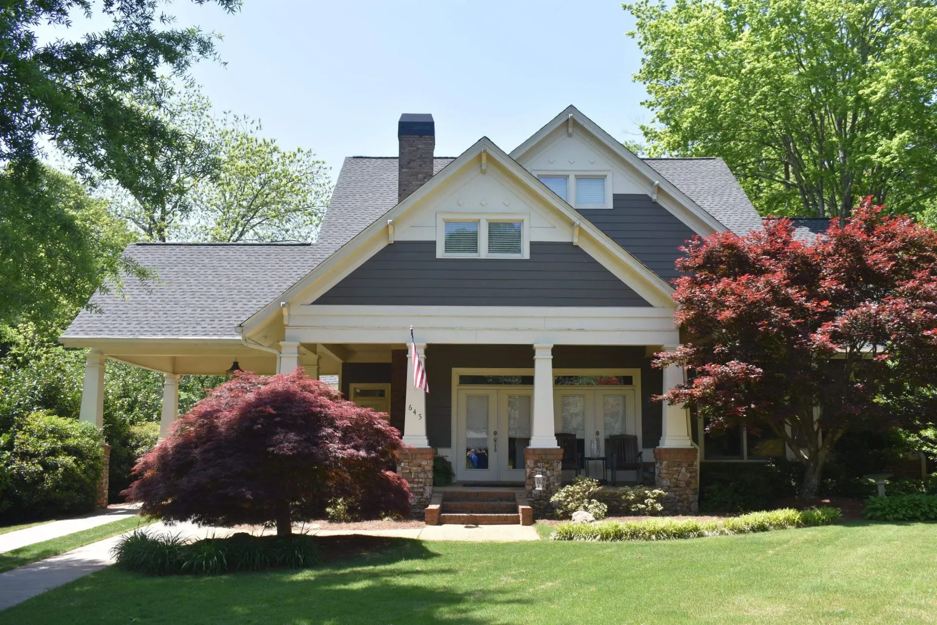Curb-side view of a Craftsman-style house with a porch, green lawn, and two trees with red leaves.