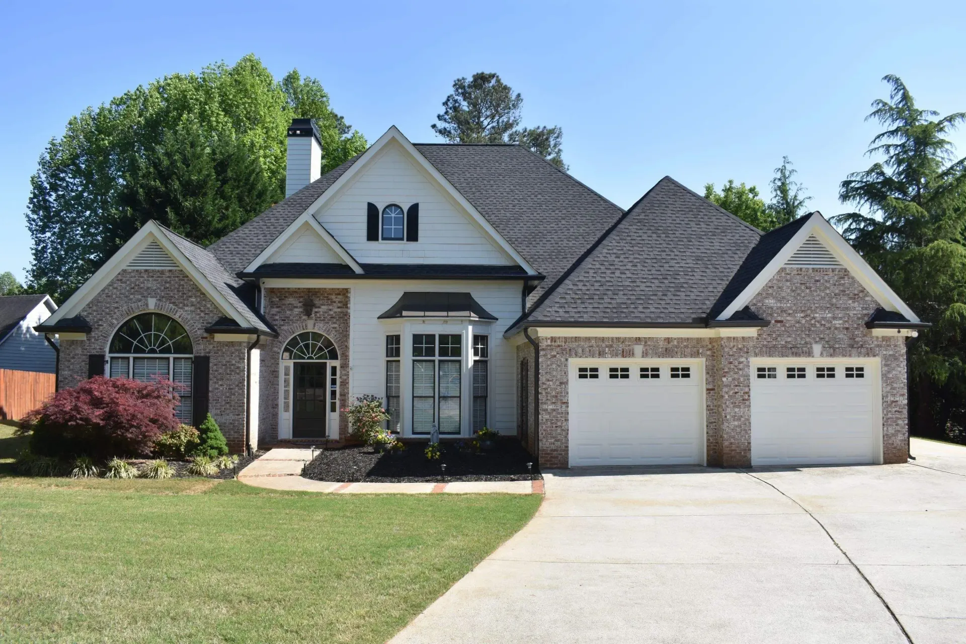 House with white and brick facade, black roof, and three-car garage, on a green lawn.