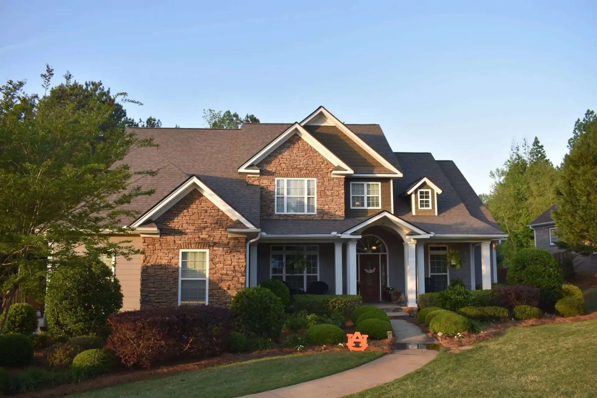 Brick house with a porch, dormers, and well-kept landscaping under a blue sky.
