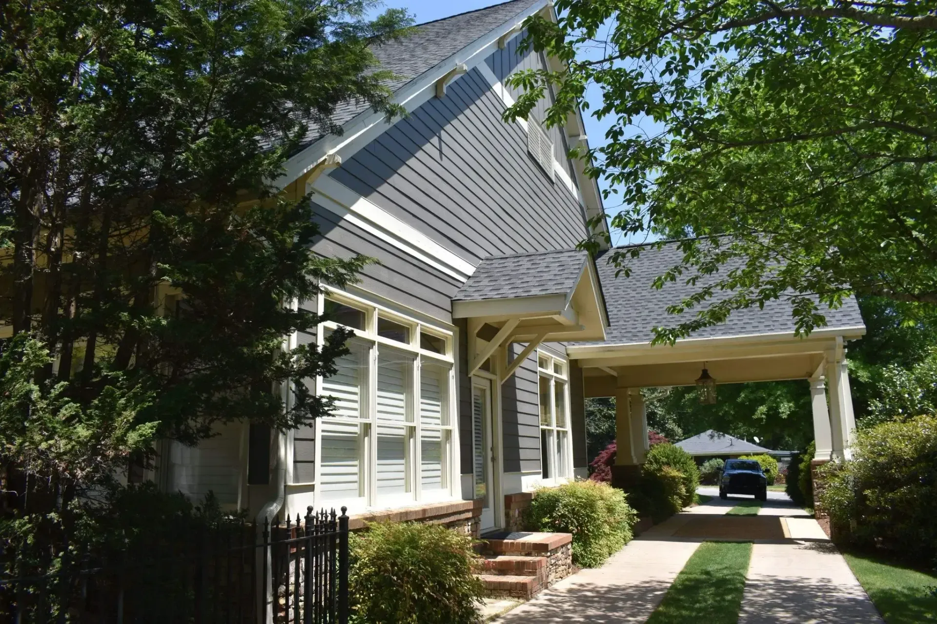 Gray house with a covered carport and a driveway lined with green turf, visible behind bushes and trees.
