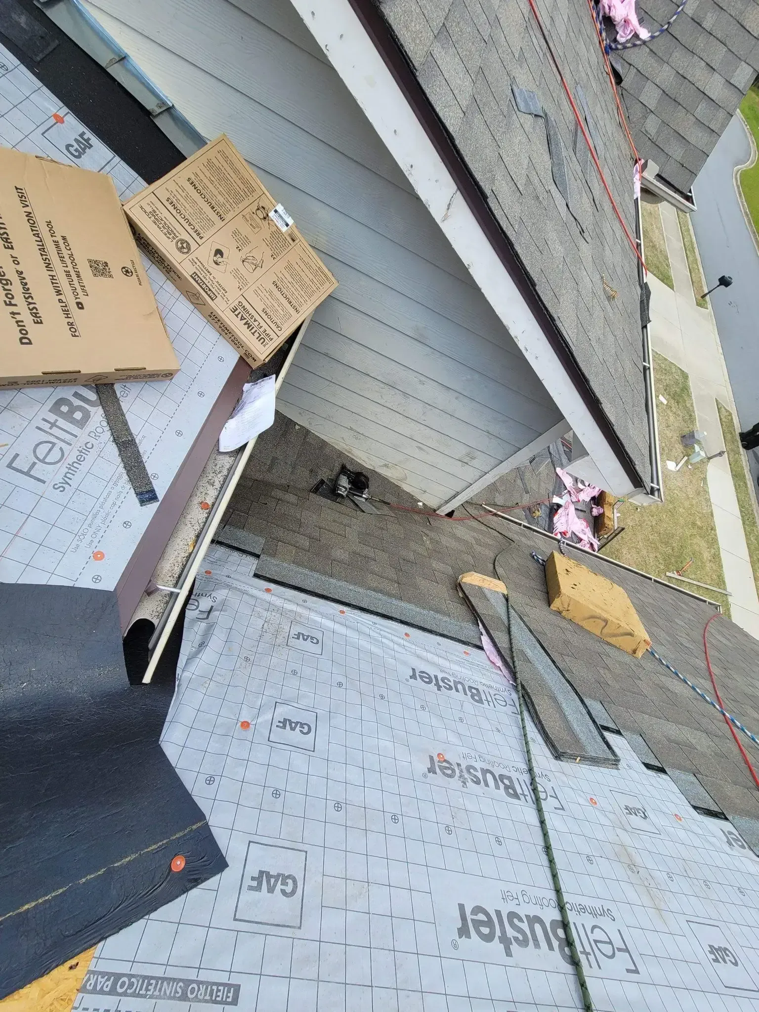 Roof partially covered with shingles, felt paper, and boxes. Brown roof with white siding.