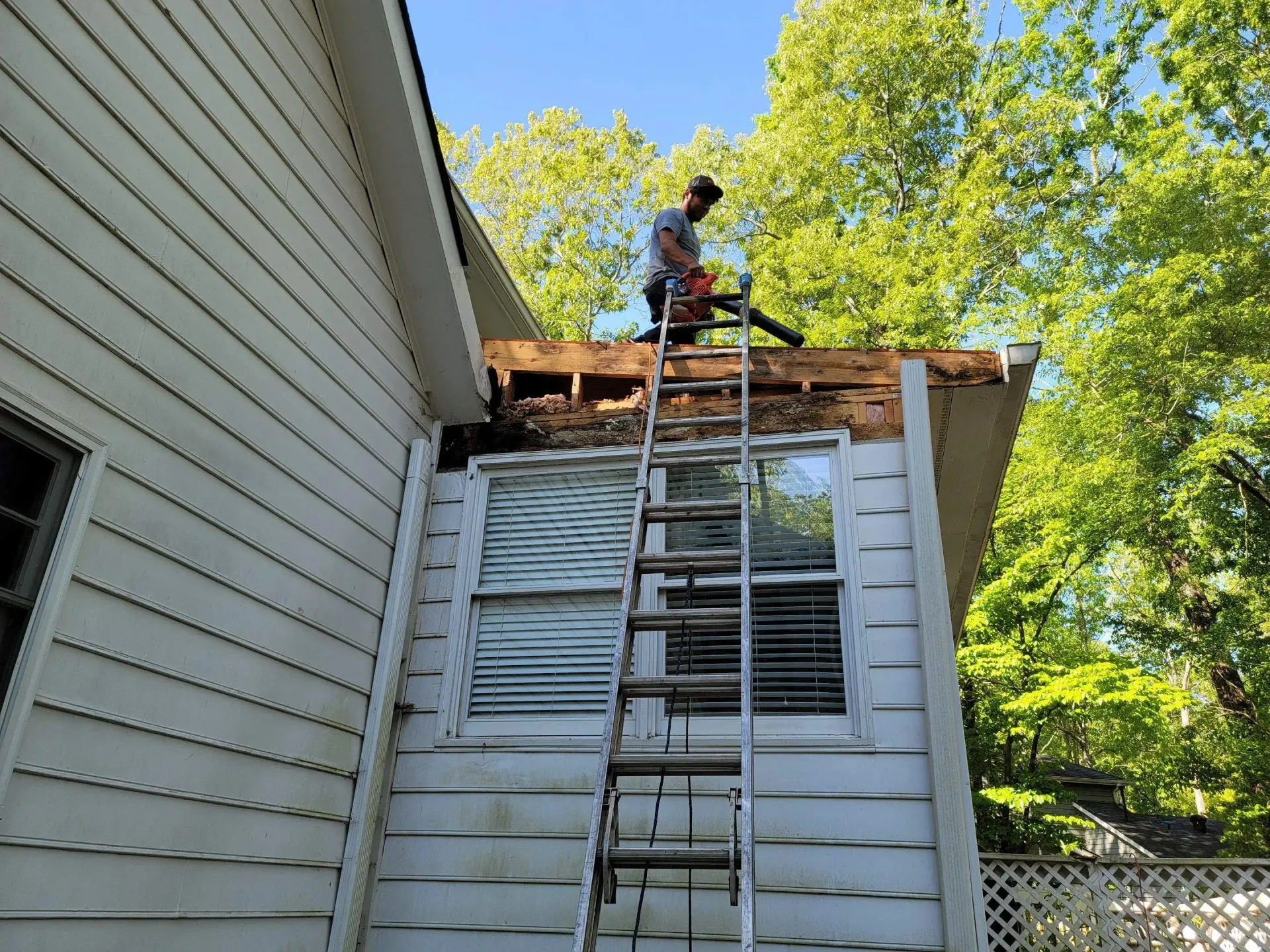 Man on ladder blowing debris from roof overhang. Building exterior, trees in background.