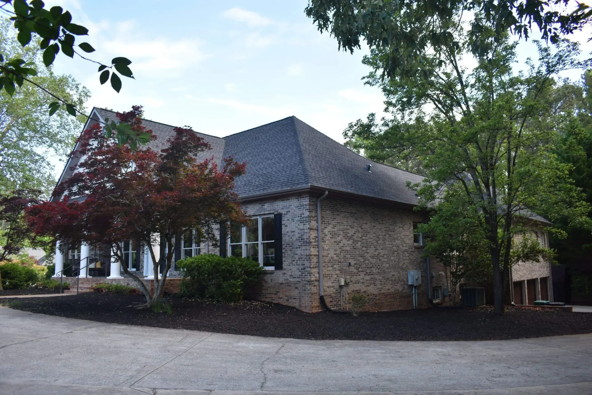 Exterior of a brick building with a dark roof and black shutters; surrounded by trees and dark mulch.