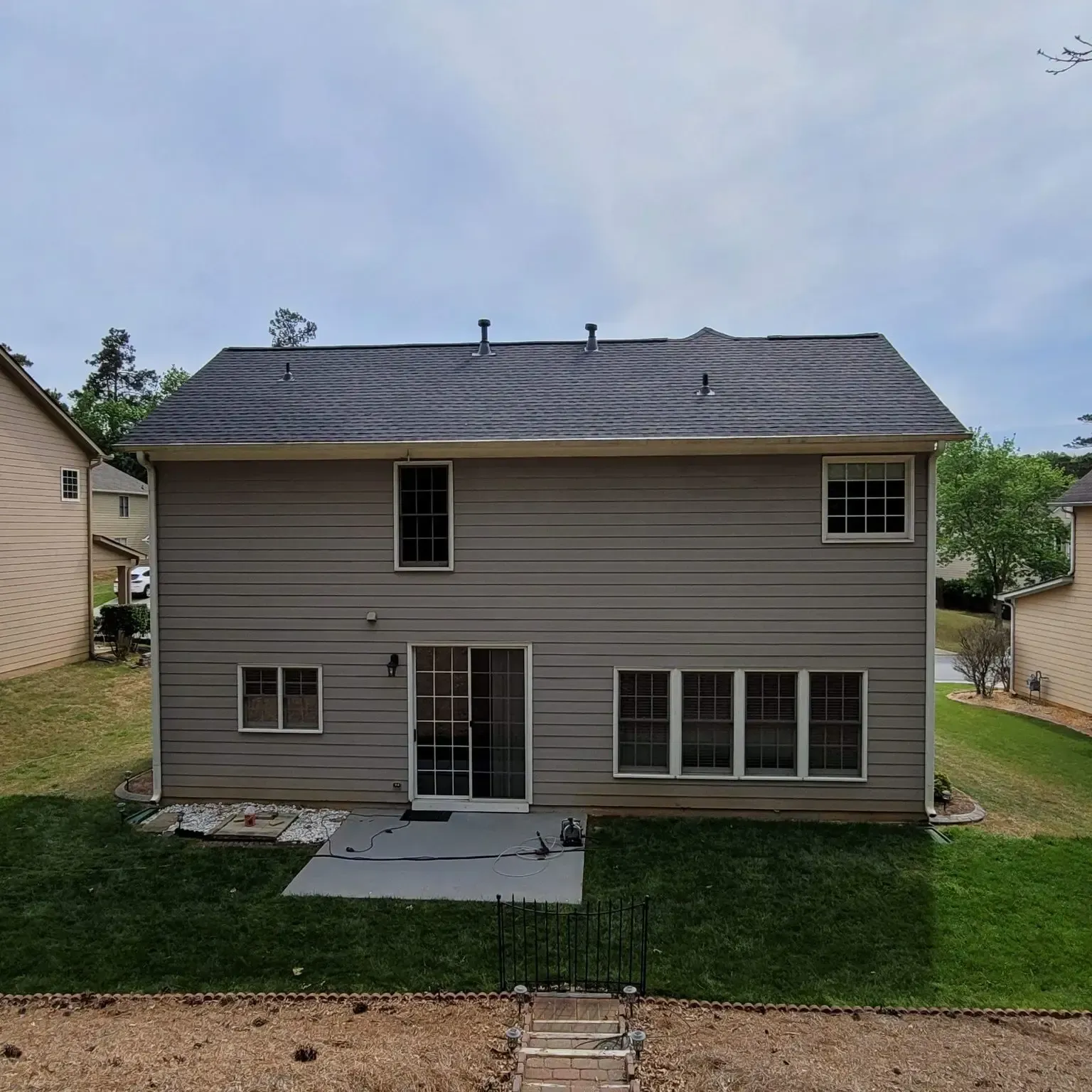Back of a two-story beige house with dark roof and multiple windows and sliding glass door. A small concrete patio in front.