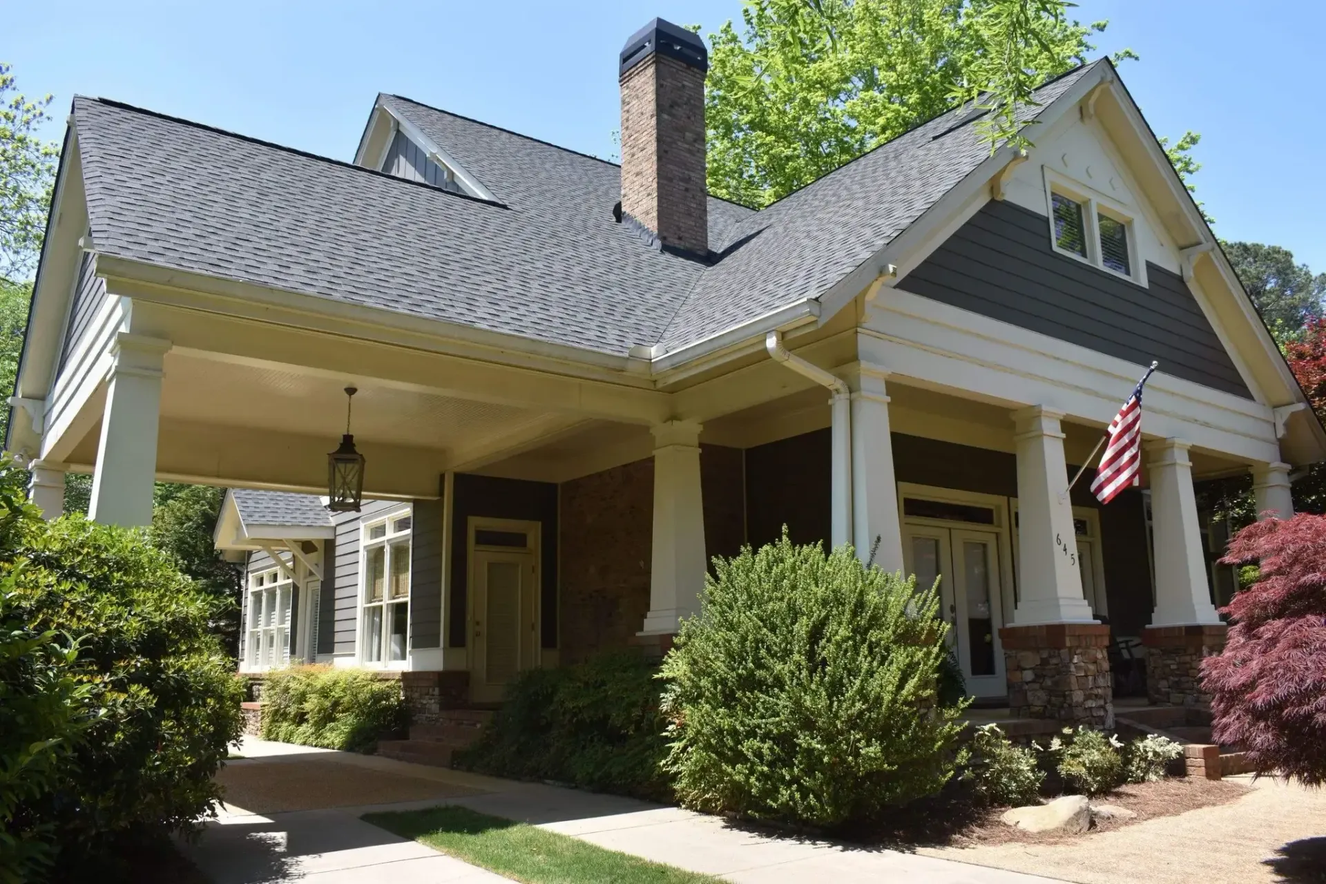 Craftsman-style home with a covered porch and brick chimney. Grey siding, white trim, and American flag.