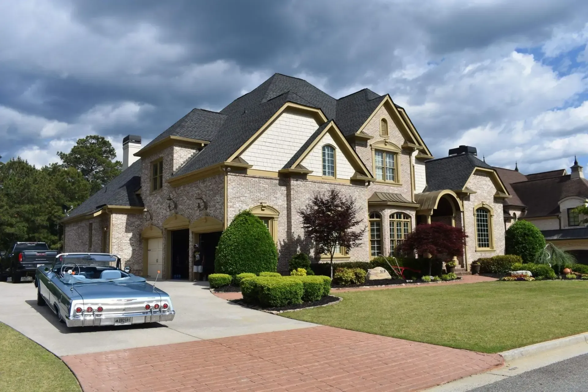 Luxury house with a blue convertible parked on a brick driveway; cloudy sky.