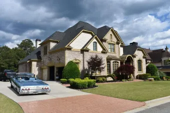 Two-story beige house with a brick driveway and a blue convertible parked in front.