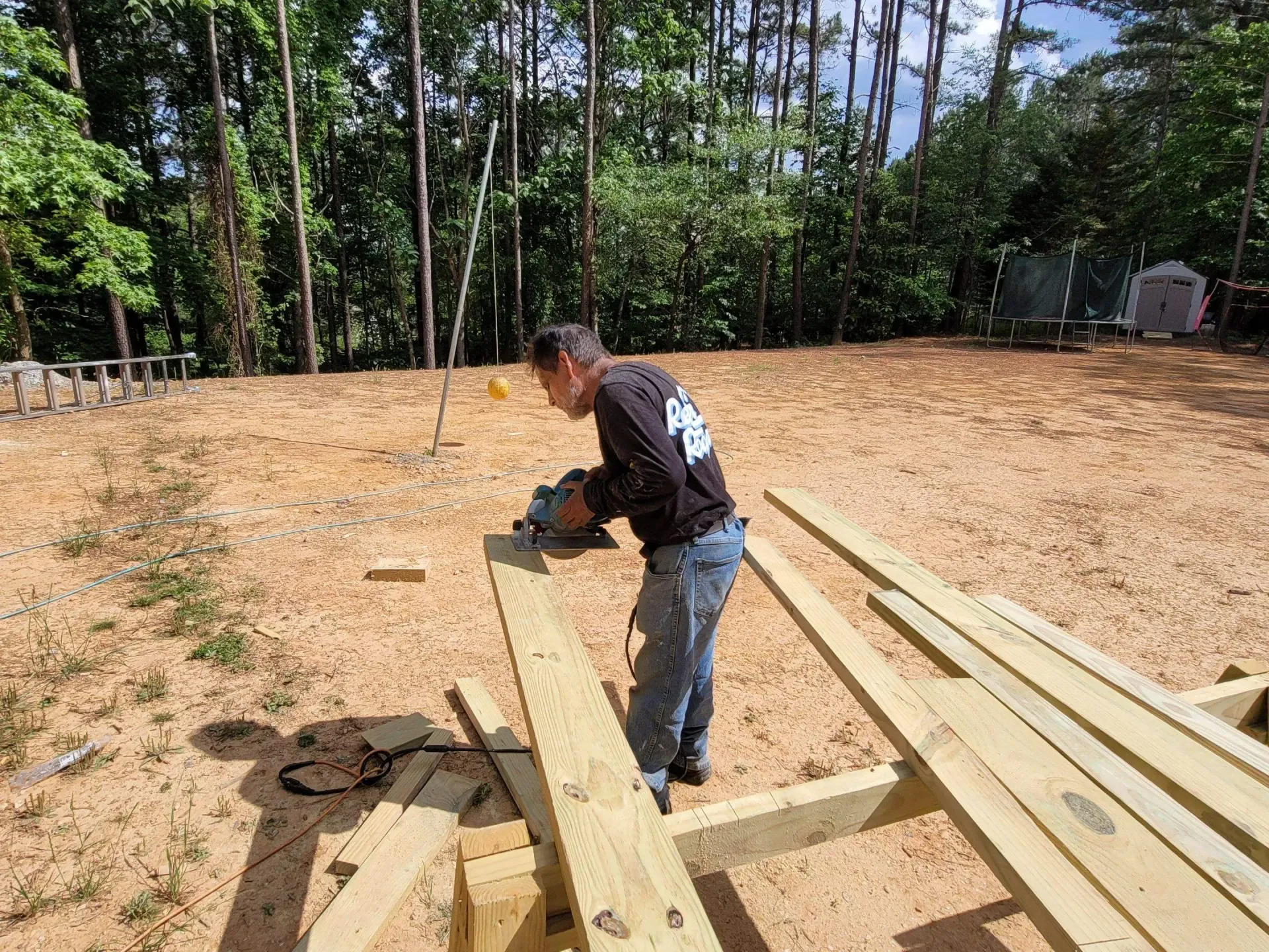 Man cuts lumber with a circular saw outdoors, constructing a deck on a sandy ground.