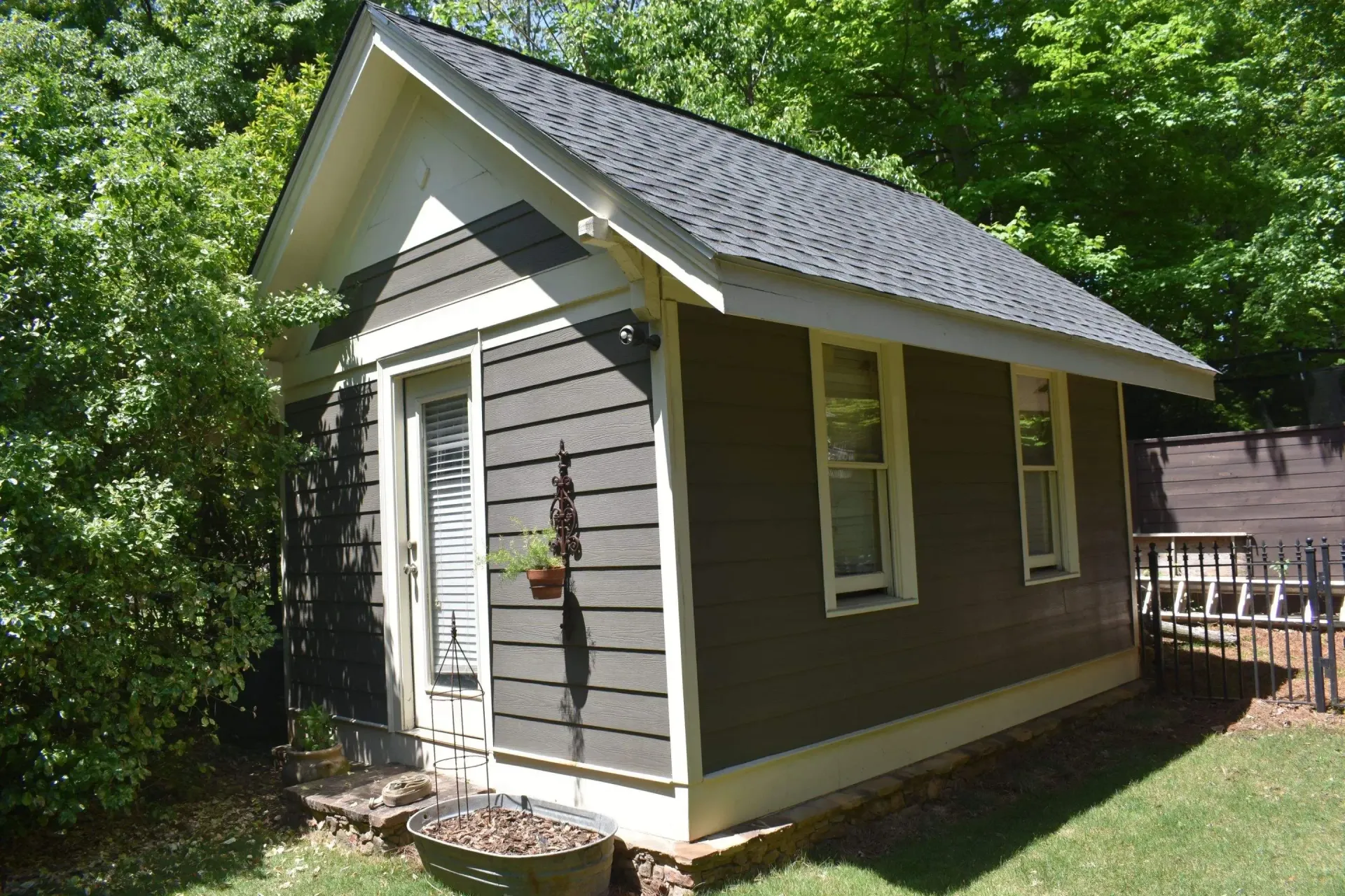 Small, brown-sided shed with white trim, a dark gray roof, and a hanging plant near the door, situated in a yard.