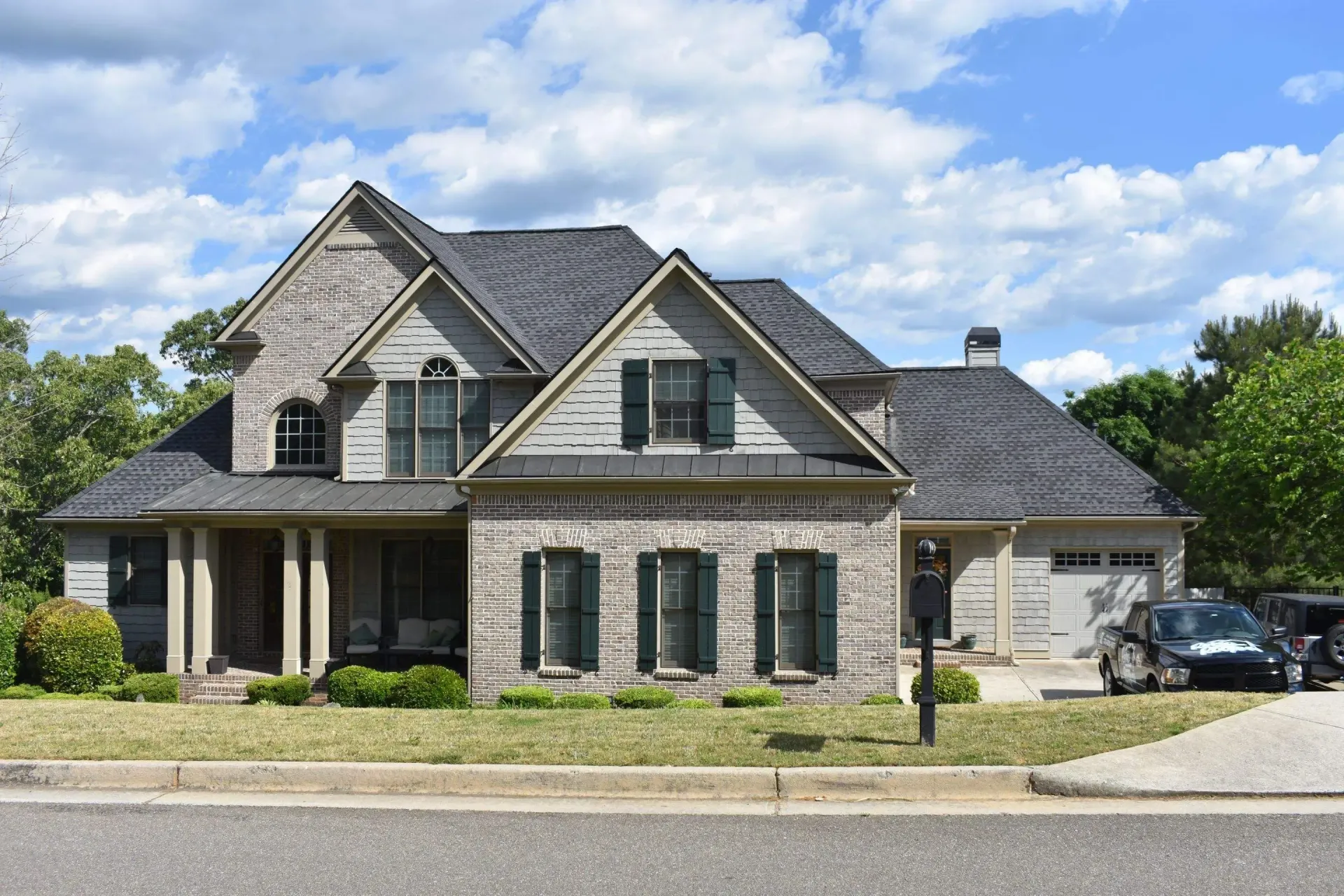 Two-story brick house with dark roof, porch, and a two-car garage under a blue sky with clouds.