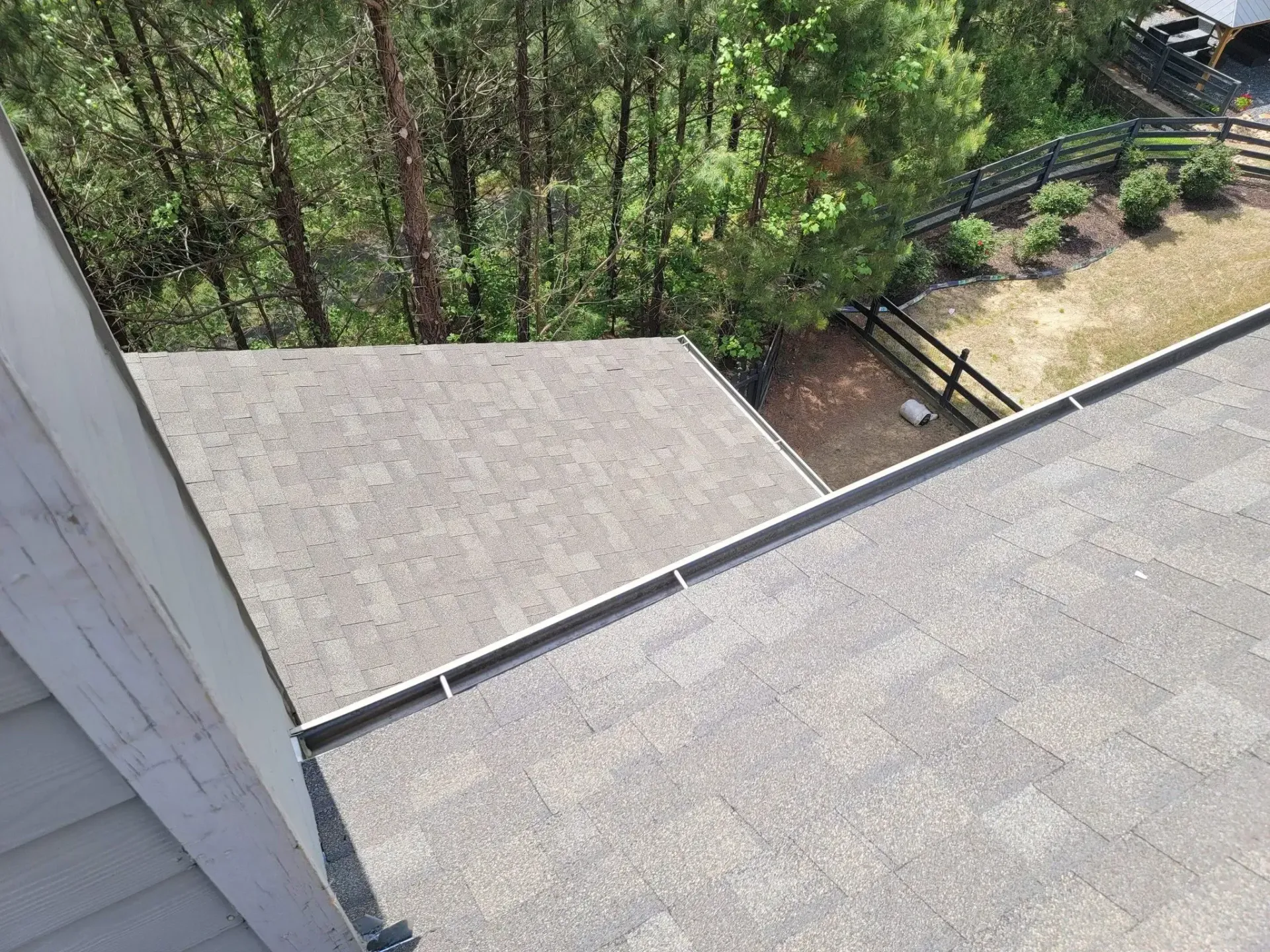 Overhead view of a gray shingle roof with a silver edge, overlooking trees and a fence.