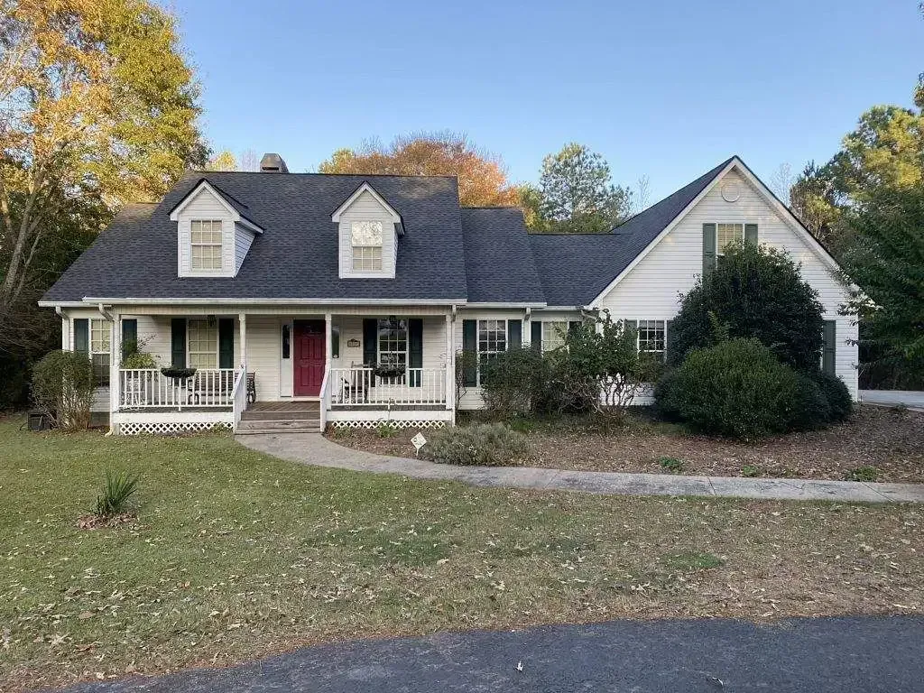 White house with dark roof and green shutters, porch, red door, and dormer windows.