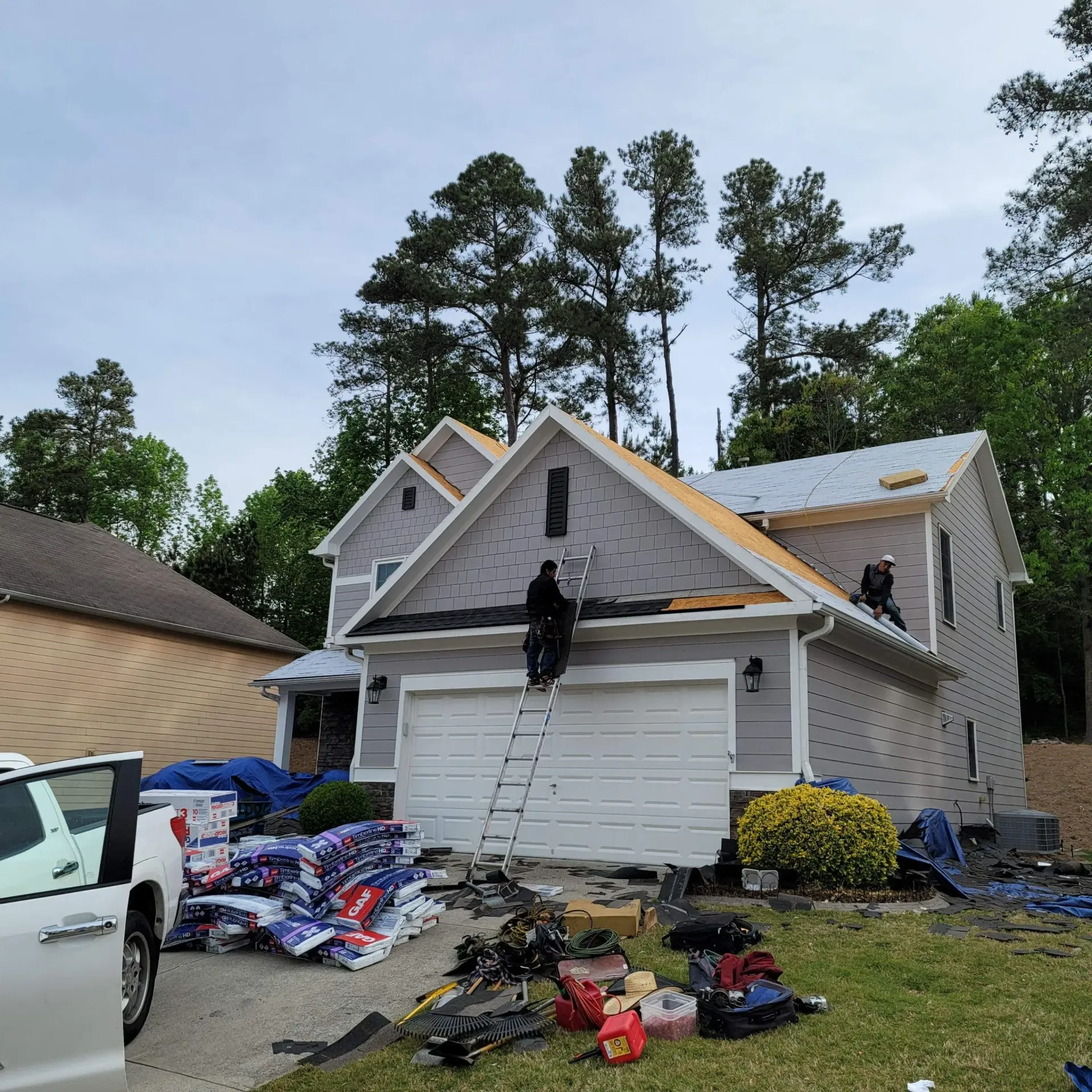 Roofers working on a two-story house, shingles partially installed. Equipment and materials are scattered on the ground.