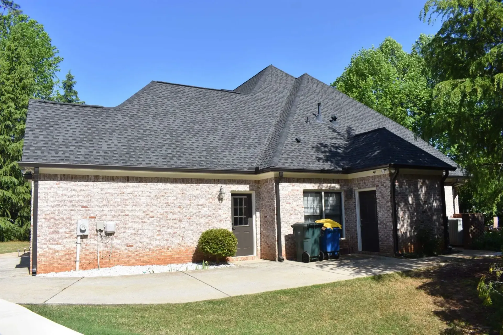 Tan brick house with gray roof, door, windows, and two trash cans. Green trees in the background.