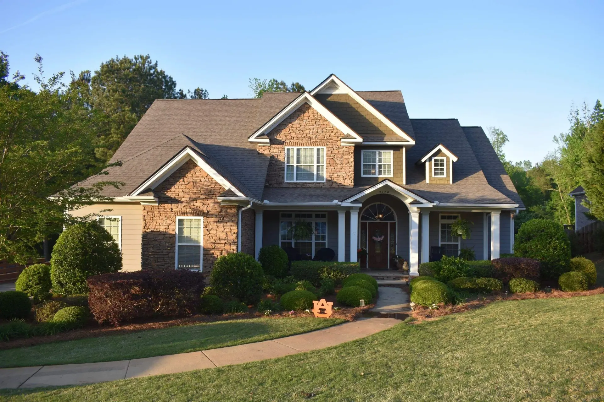 Two-story house with stone and gray siding, porch, and well-manicured landscaping in a sunny setting.
