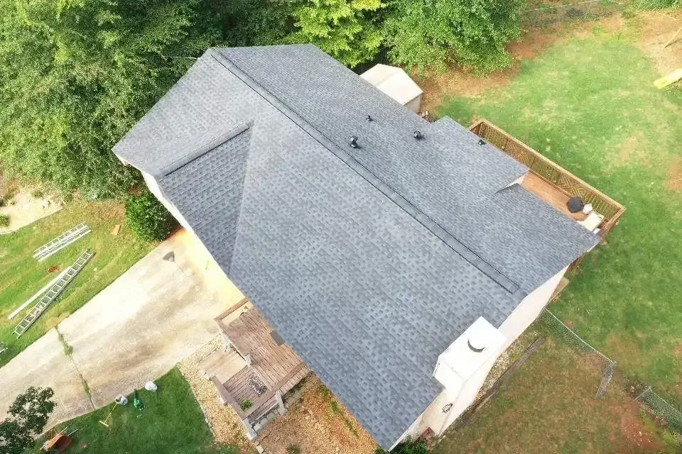 Aerial view of a house with a dark gray shingle roof, surrounded by green trees and grass.