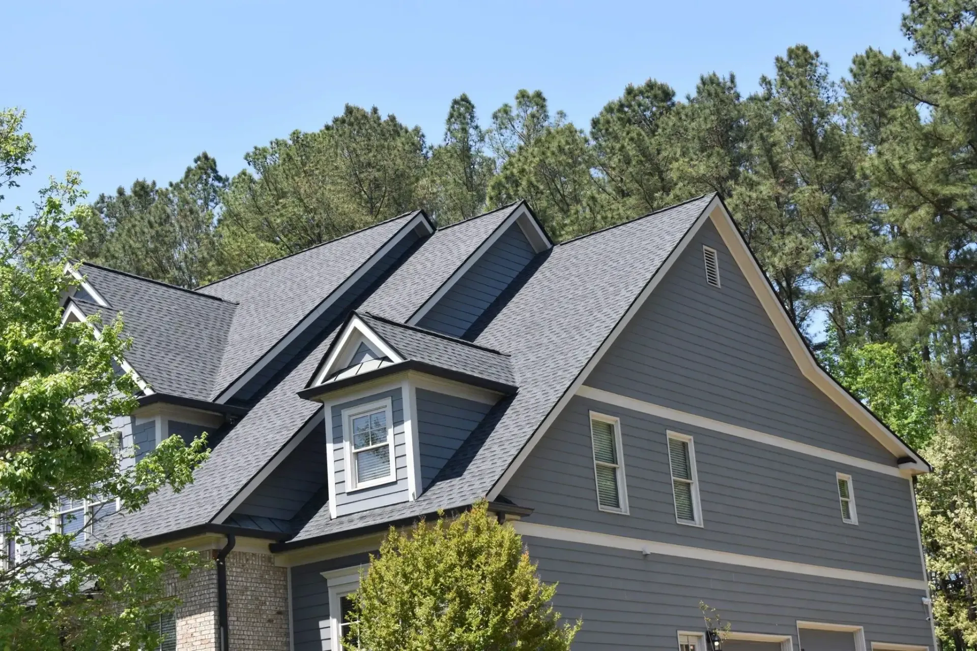 Gray house with dark gray shingle roof and blue siding against a background of green trees and blue sky.