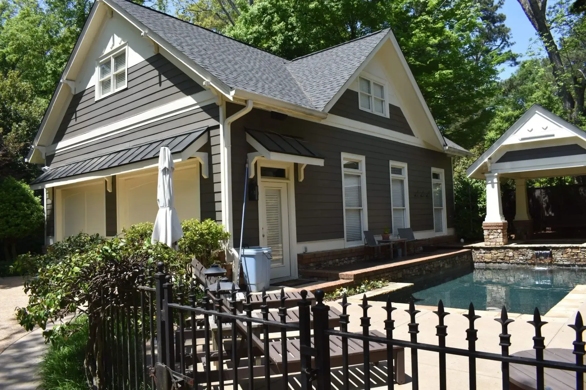 Dark gray house with white trim, near a pool and black fence, surrounded by greenery.
