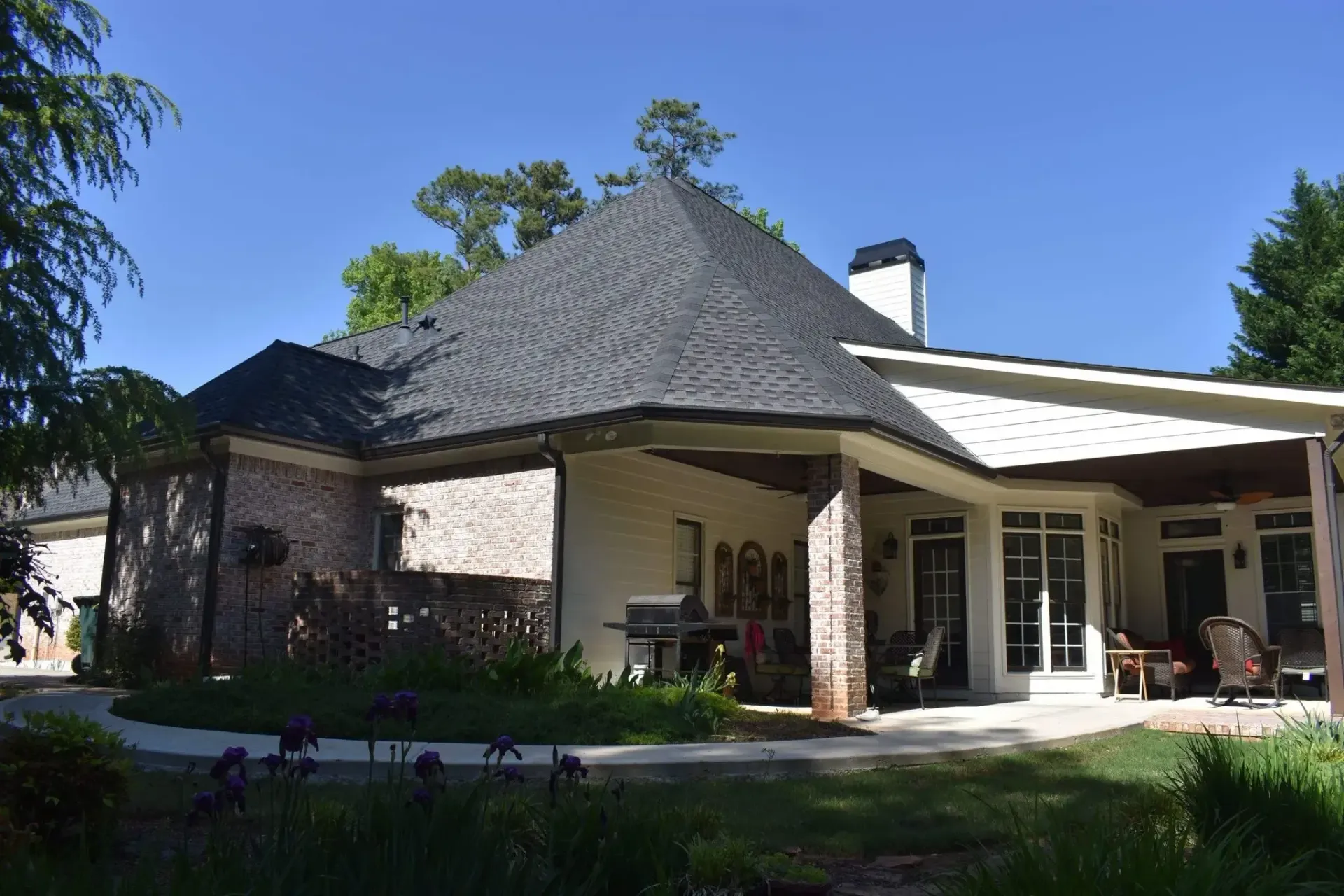 Rear view of a light-colored brick and white house with a dark roof, covered patio, and green yard.