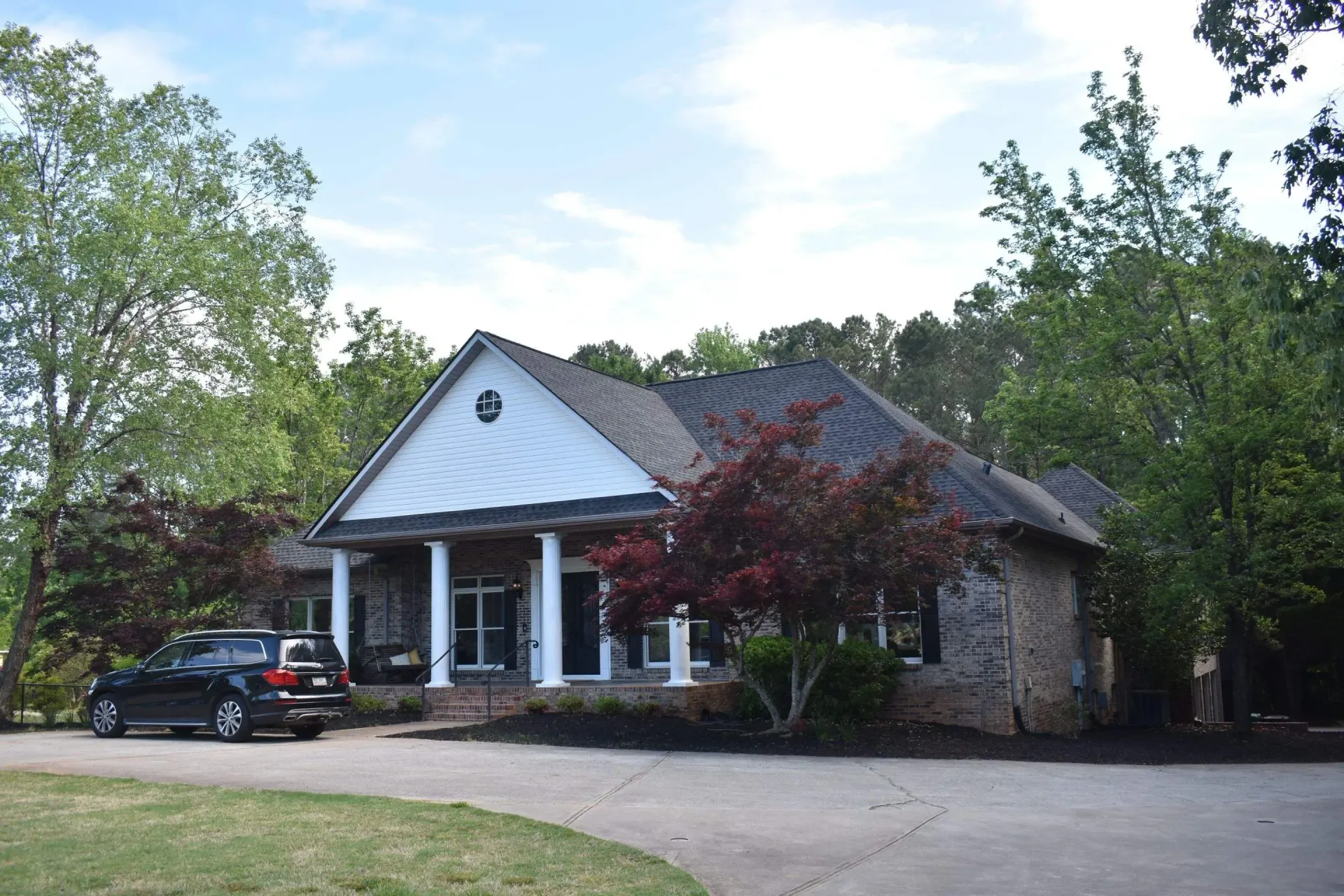 A one-story brick house with a white columned porch, black car in the driveway, and surrounded by trees.