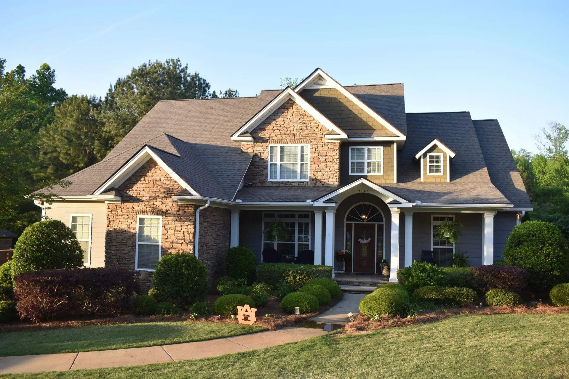 Two-story house with brick and stone exterior, green lawn, and landscaped bushes.