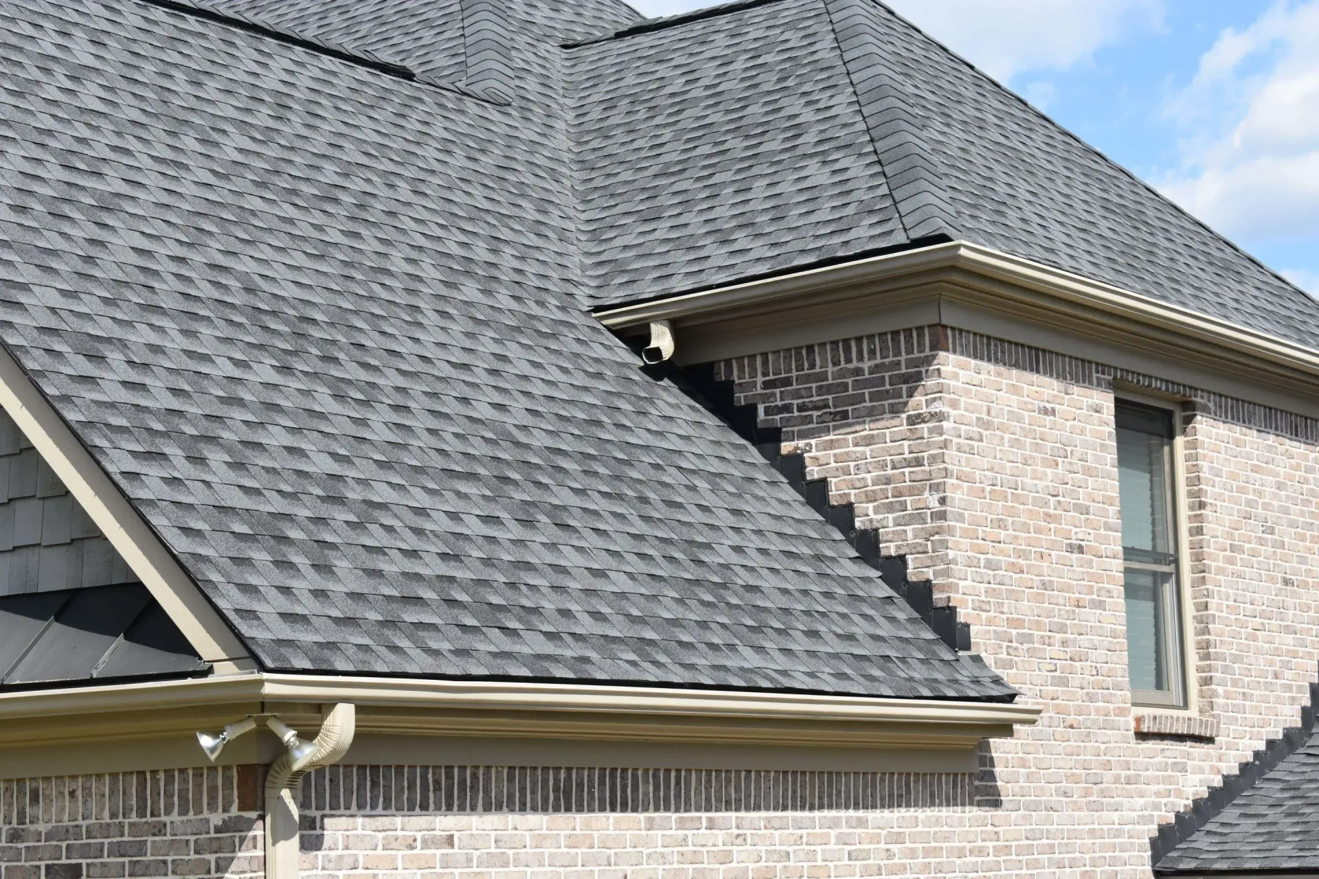 Gray asphalt shingle roof on a brick house with tan gutters. Sunny day.