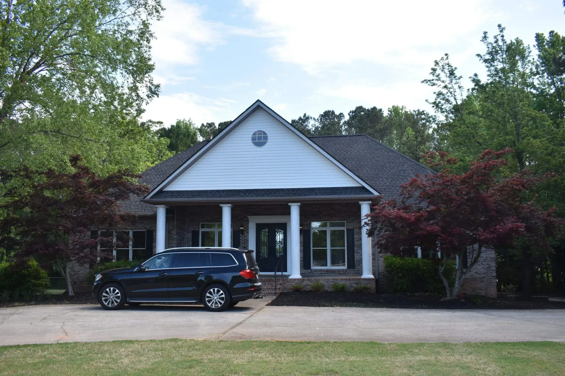 Black SUV parked in front of a one-story house with white columns, trees, and a blue sky.