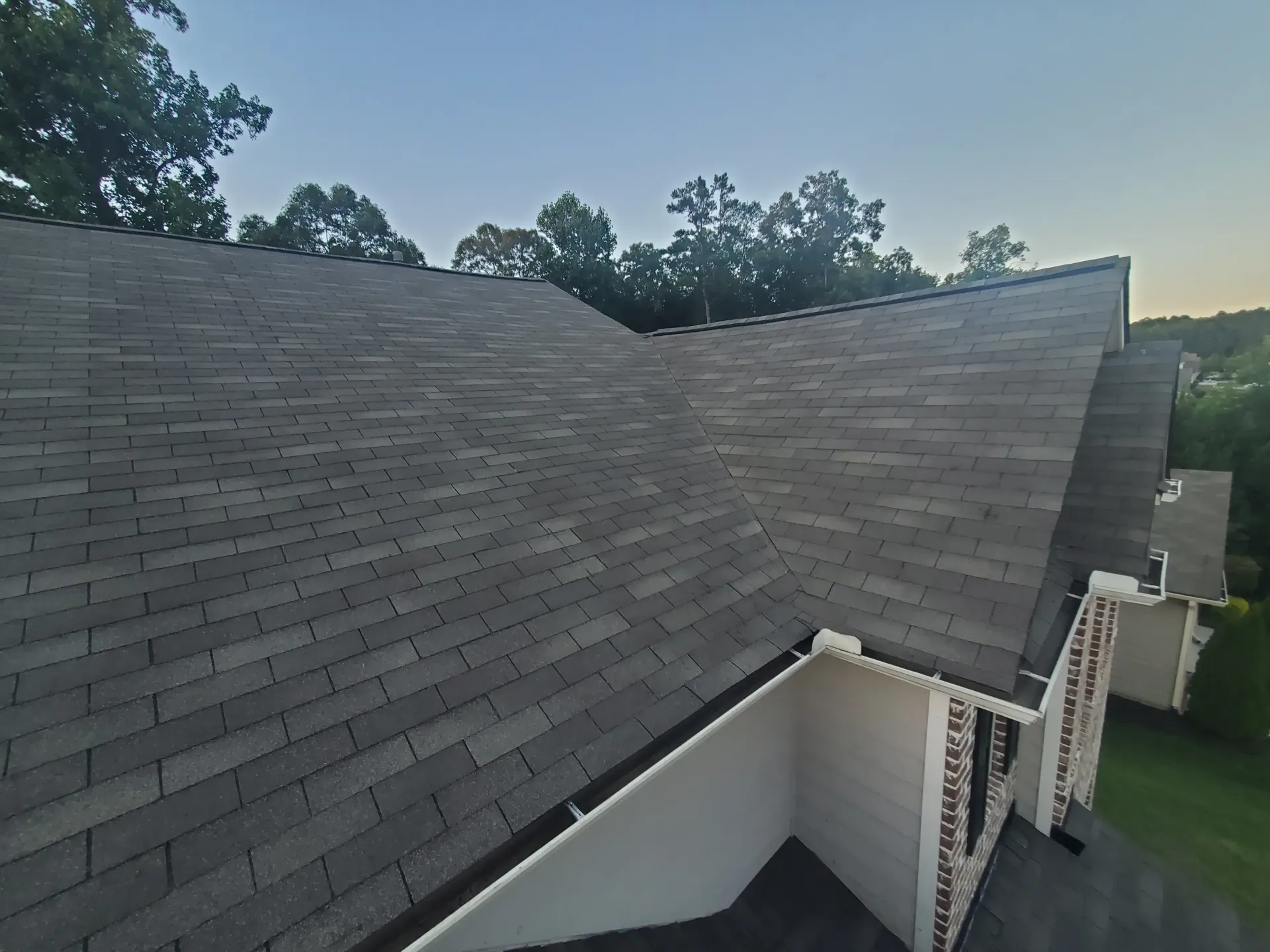 Gray shingle roof of a house with trees in the background under a blue sky.