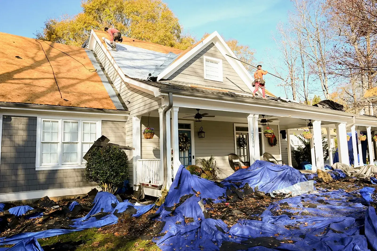 Roofers replacing shingles on a house; blue tarps on the ground, trees in the background.