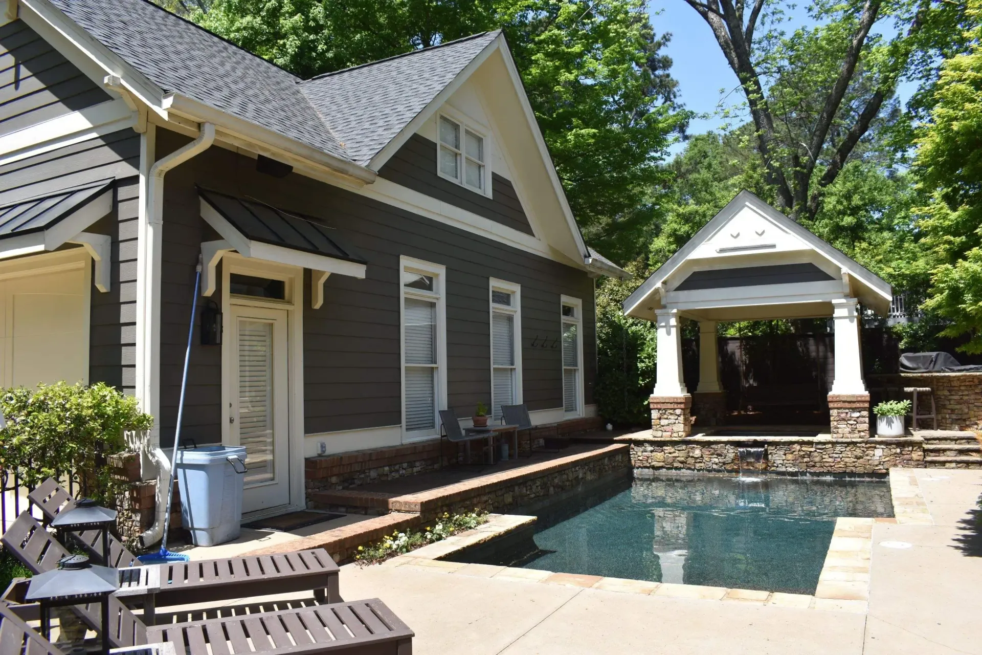 A house with a dark facade next to a pool and pavilion on a sunny day.