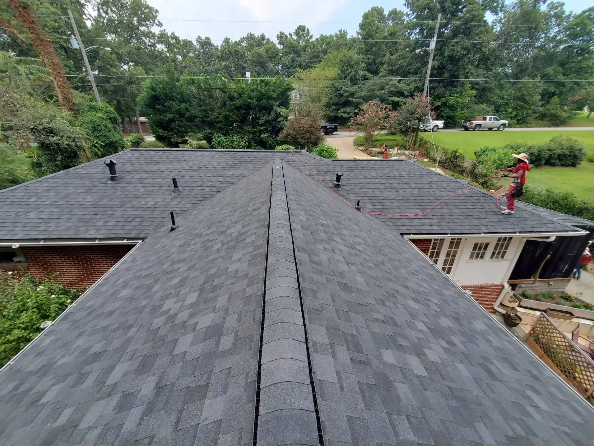 View from a roof, showing dark gray shingle roof, vents, and a person working on the side near trees.