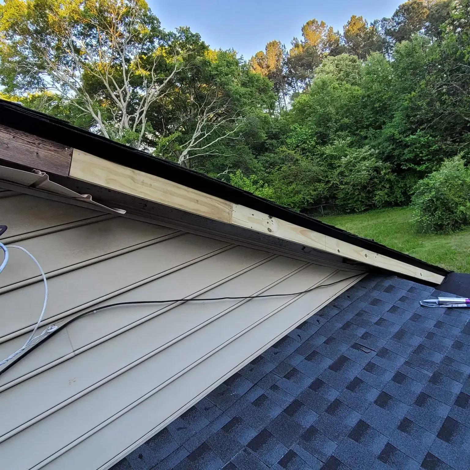 Roof repair: New wood installed on the edge of a house roof, next to vinyl siding and a shingled roof.