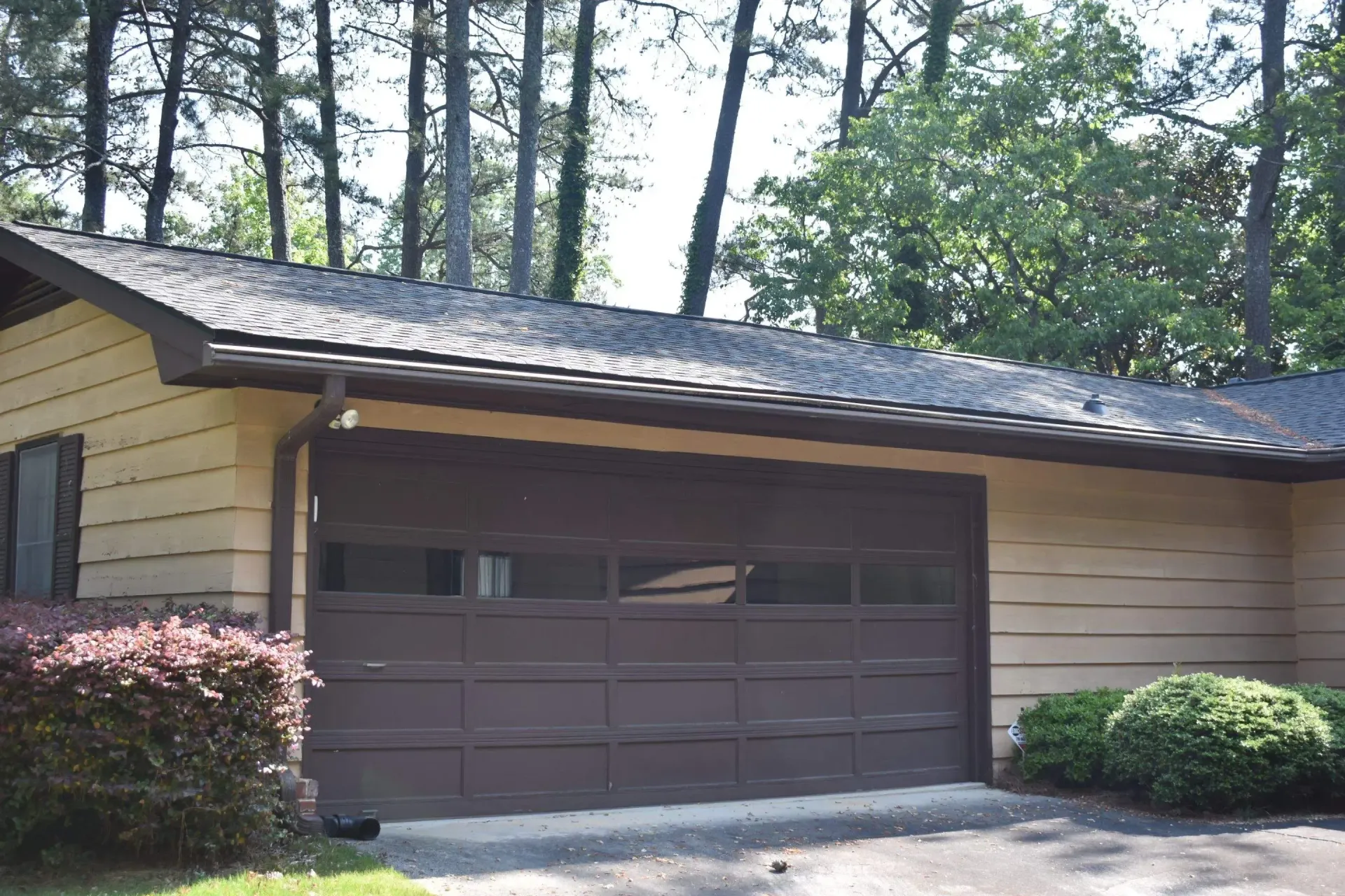 Brown garage door on a tan house with dark roof and landscaping in front of trees.