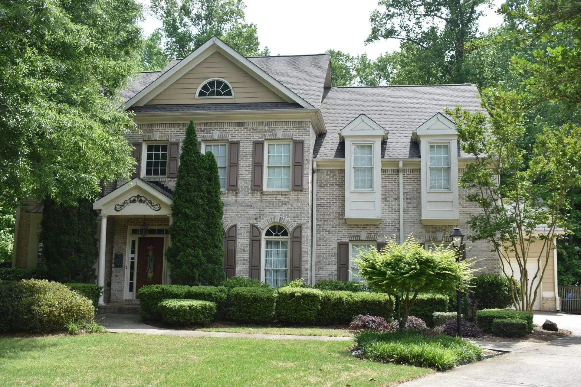 Two-story brick house with a gray roof and dormer windows, surrounded by green trees and bushes, on a sunny day.