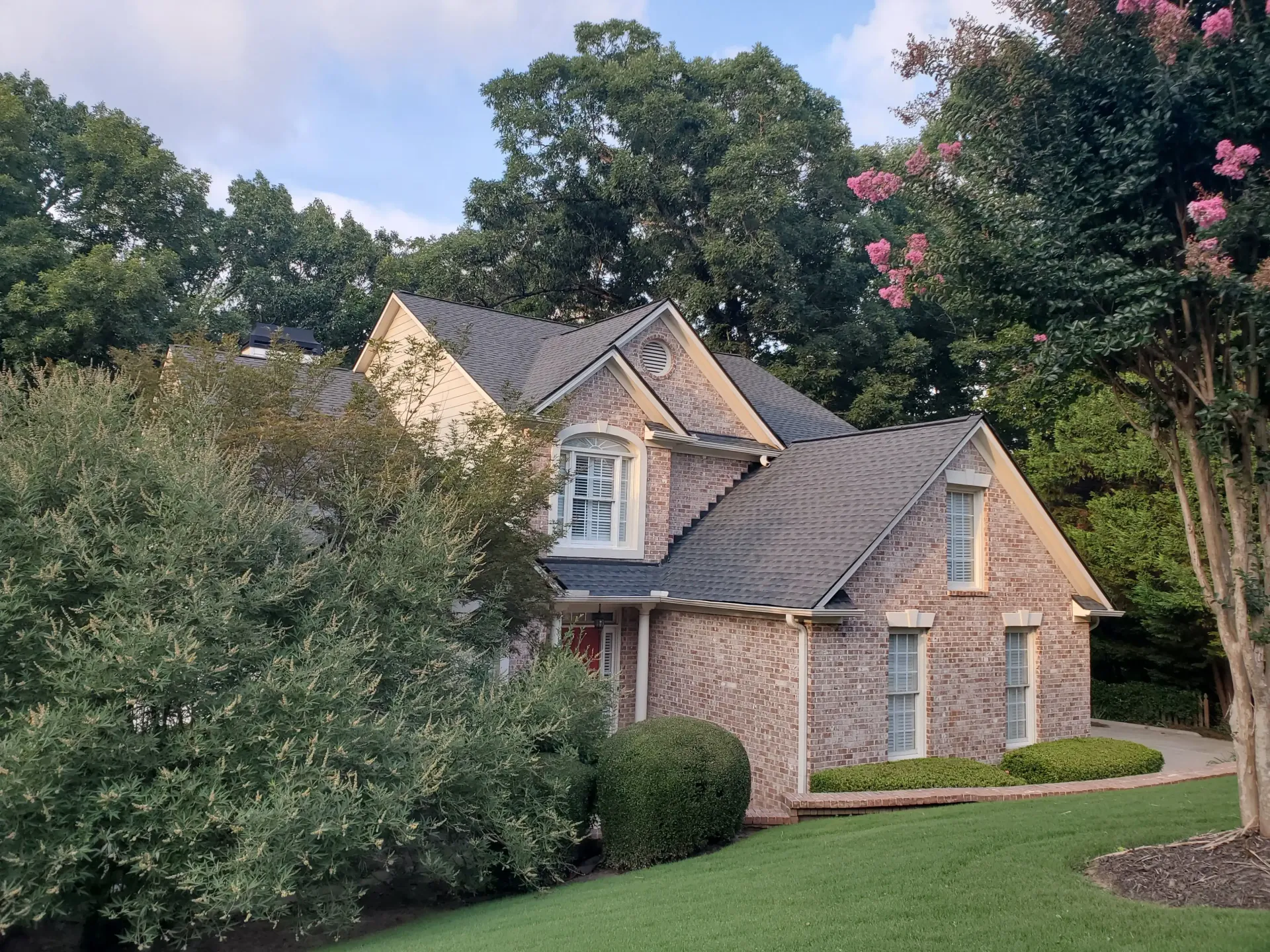 Brick house with dark roof and green lawn surrounded by trees and bushes.