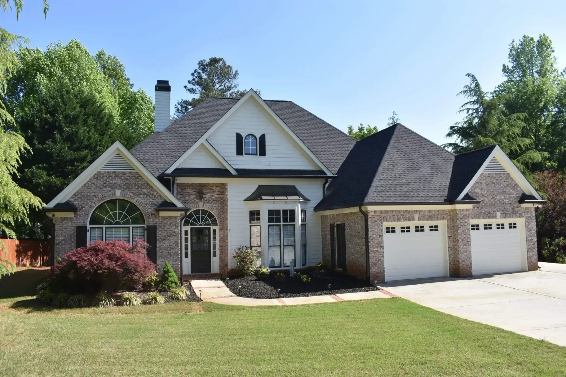 Suburban home with white siding, brick accents, black roof, three-car garage, and manicured lawn.