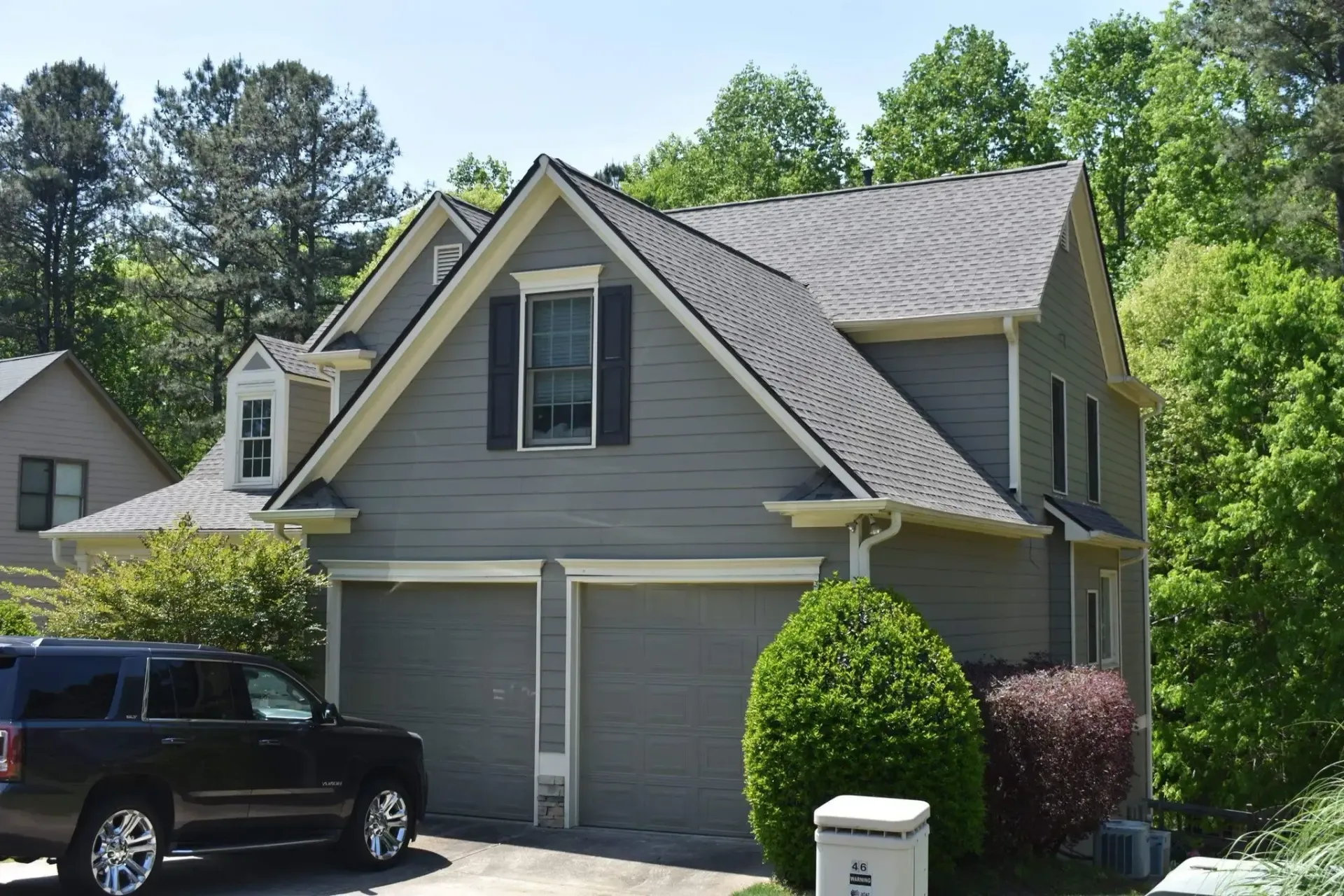 Grey two-story house with a two-car garage. A black SUV is parked in the driveway. Trees in the background.