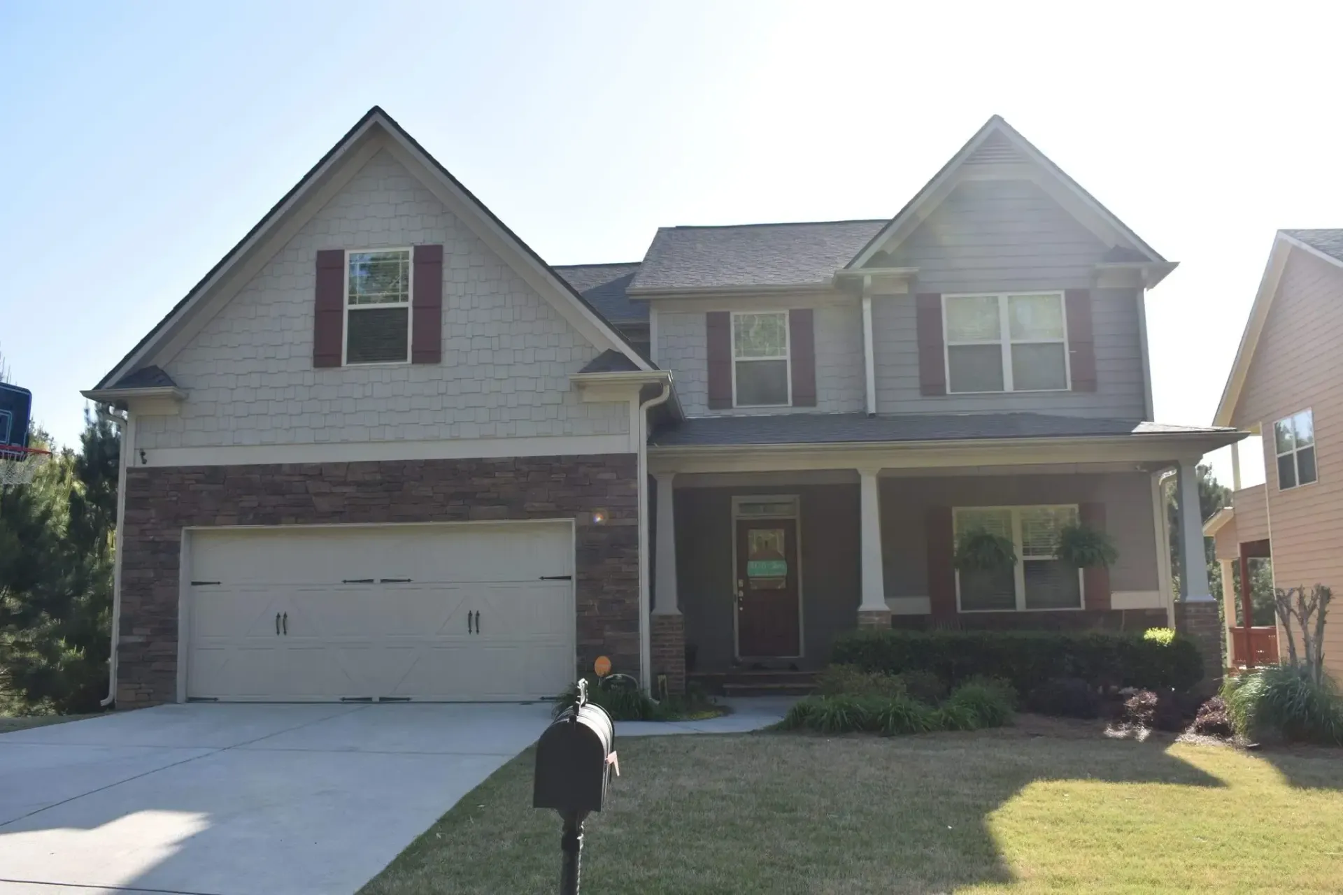 Two-story house with tan siding, brick facade, and a two-car garage. Front porch and green lawn.