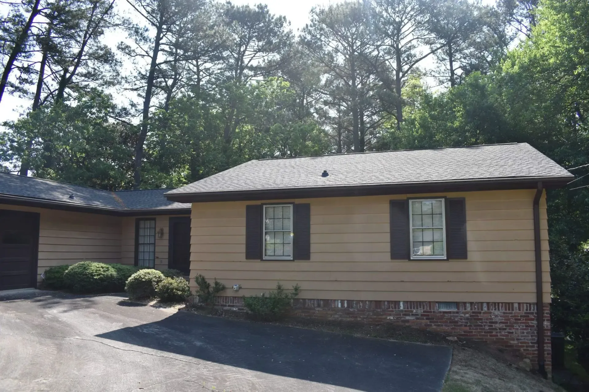 Tan house with dark shutters, brick base, and a driveway, surrounded by trees.