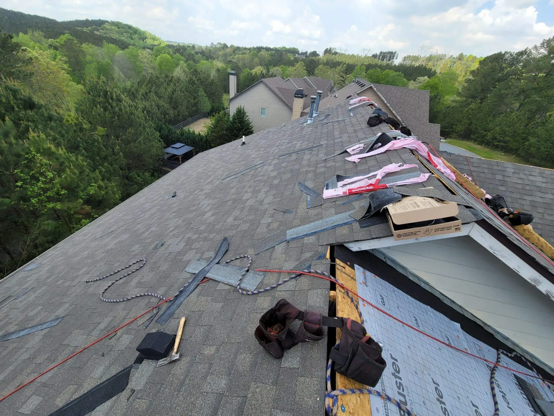 Roof being repaired, shingles partially removed. Tools and materials on the roof with trees and a house in the background.