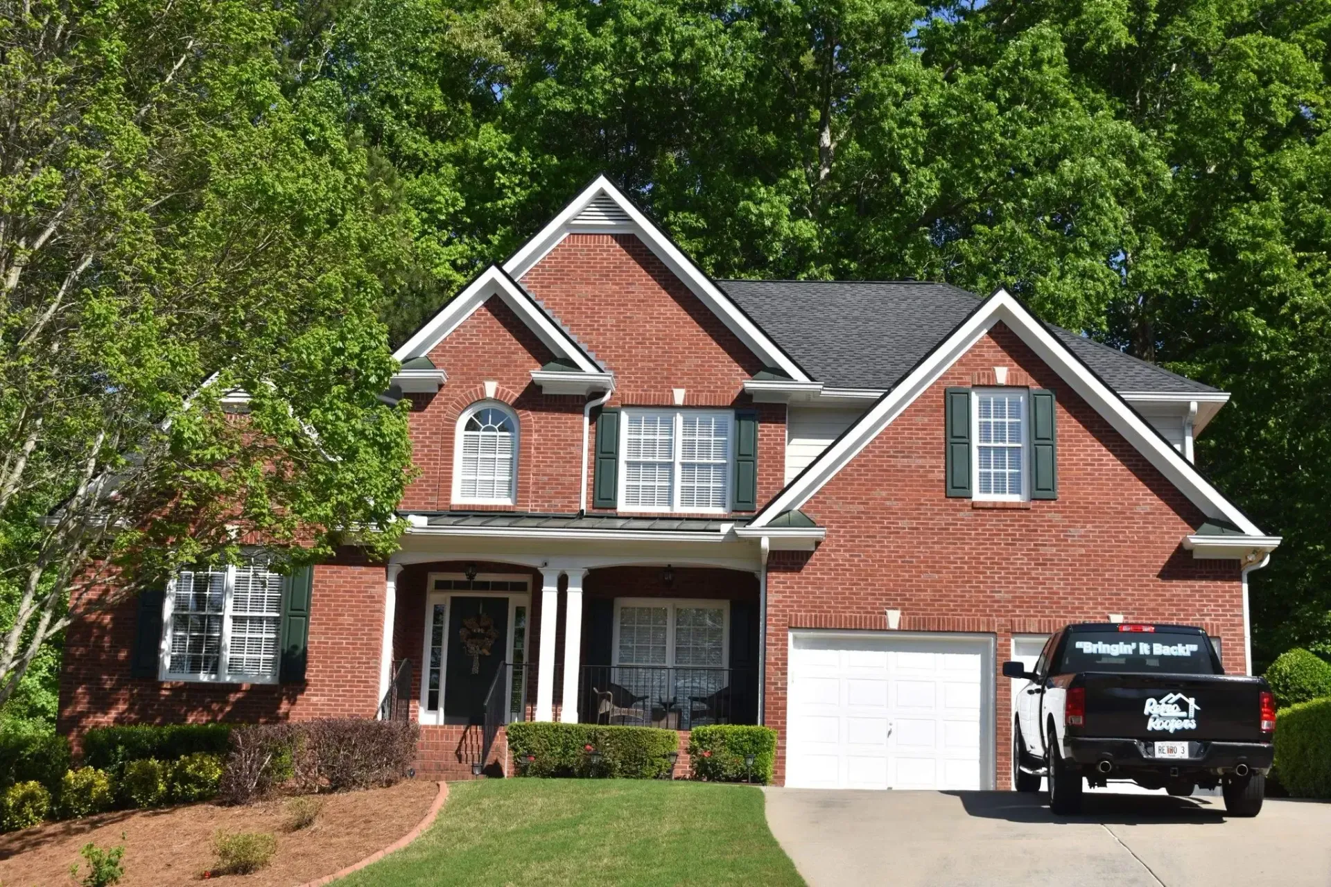 Red brick two-story house with a black pickup truck parked in the driveway; green trees in the background.