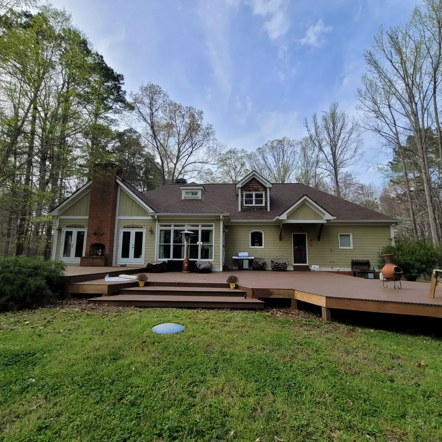 Back of a light green house with brown roof and large wooden deck on a grassy lawn.