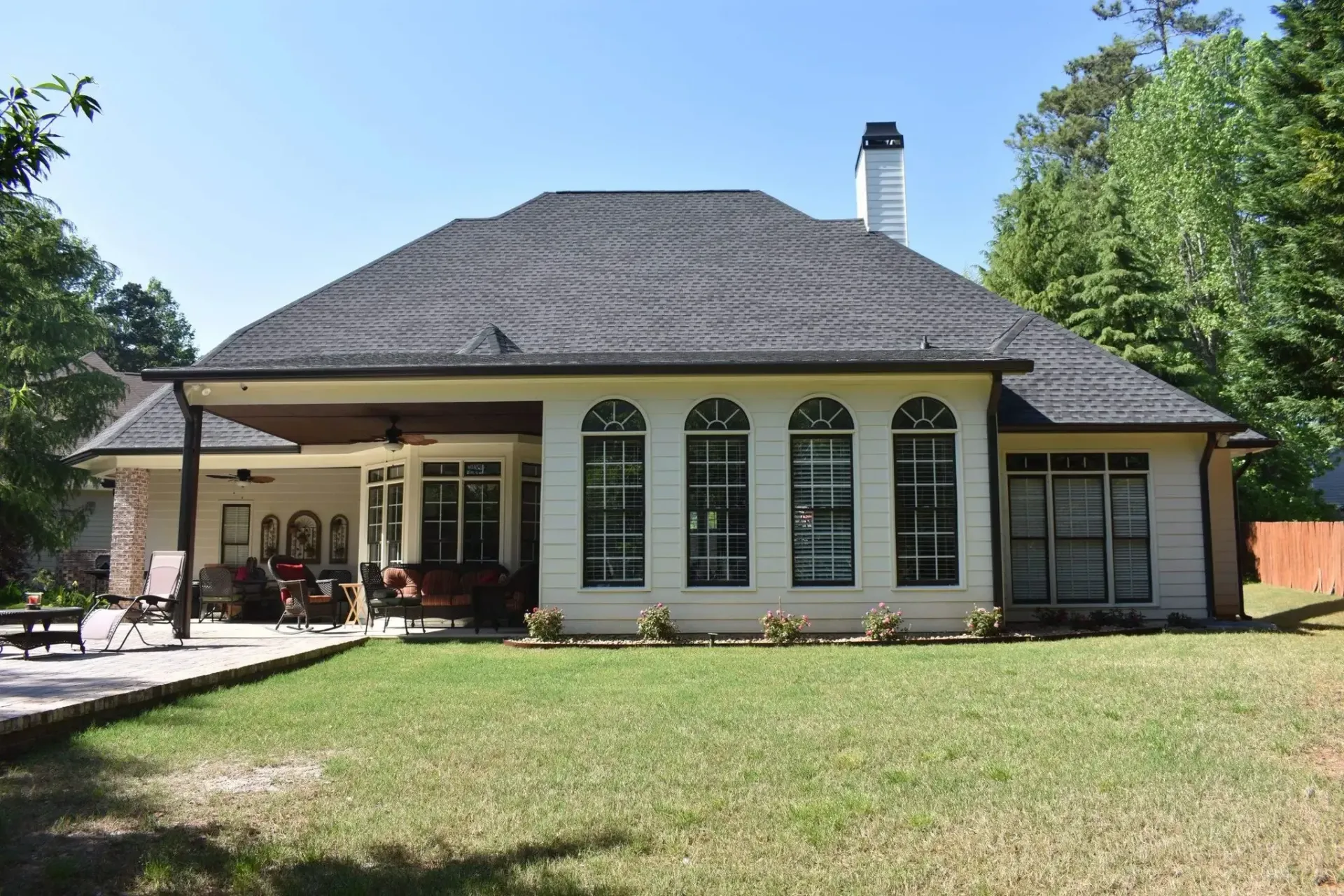 Back of a beige house with a dark roof and multiple arched windows, featuring a covered patio, on a sunny day.