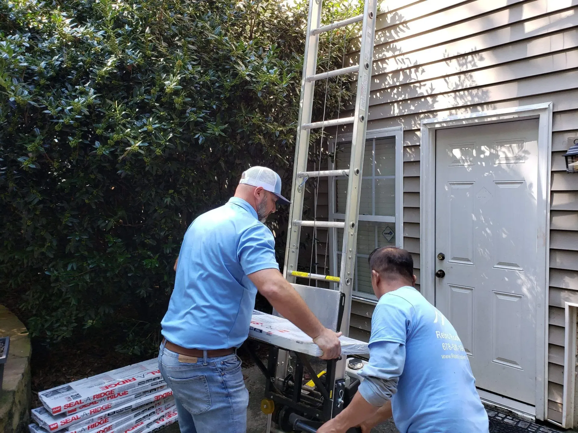 Two people in blue shirts move construction materials next to a house with a ladder.