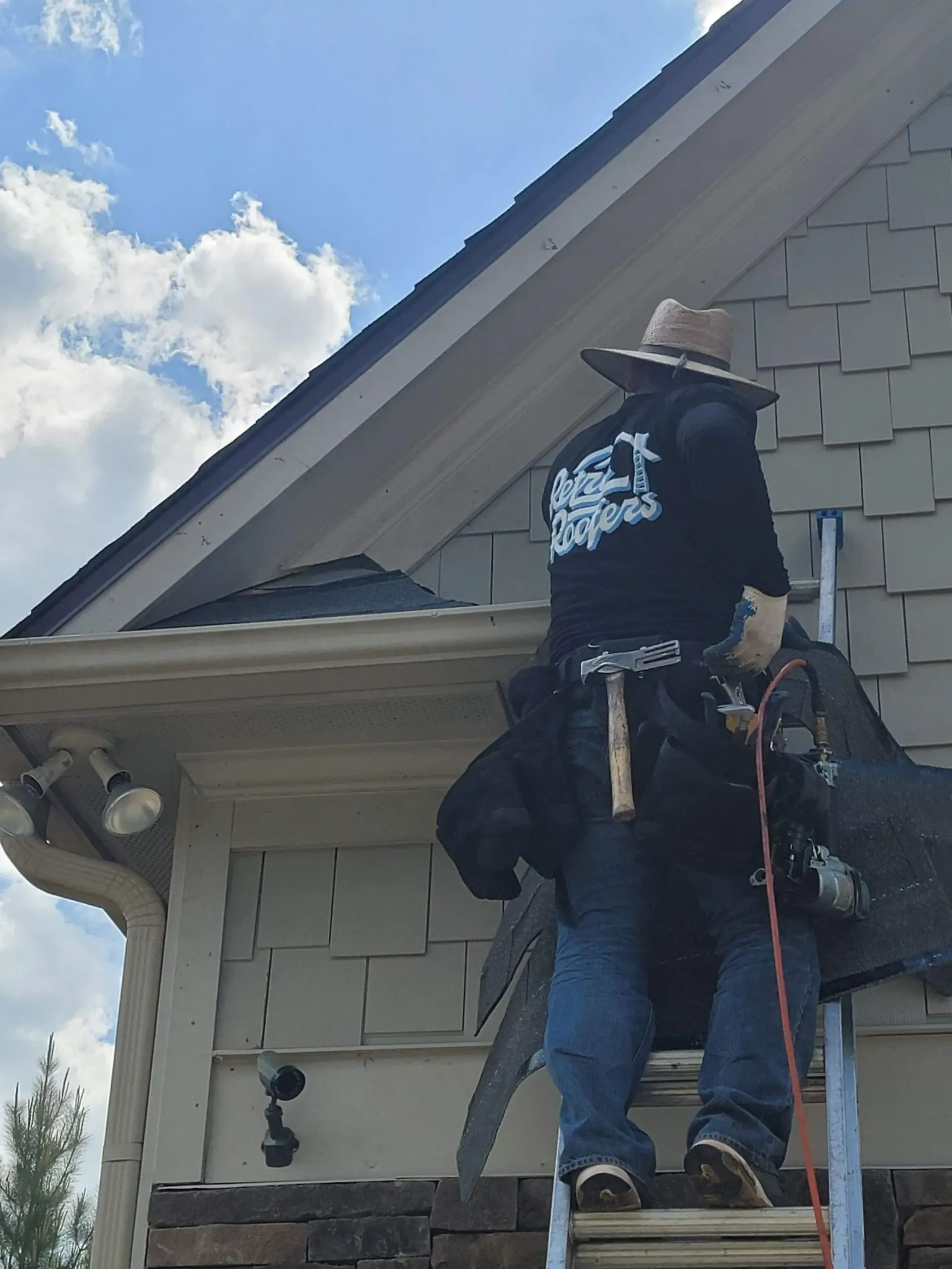 Roofer on a ladder near a house's eaves, wearing a hat, black shirt, and tool belt, working on the roof.