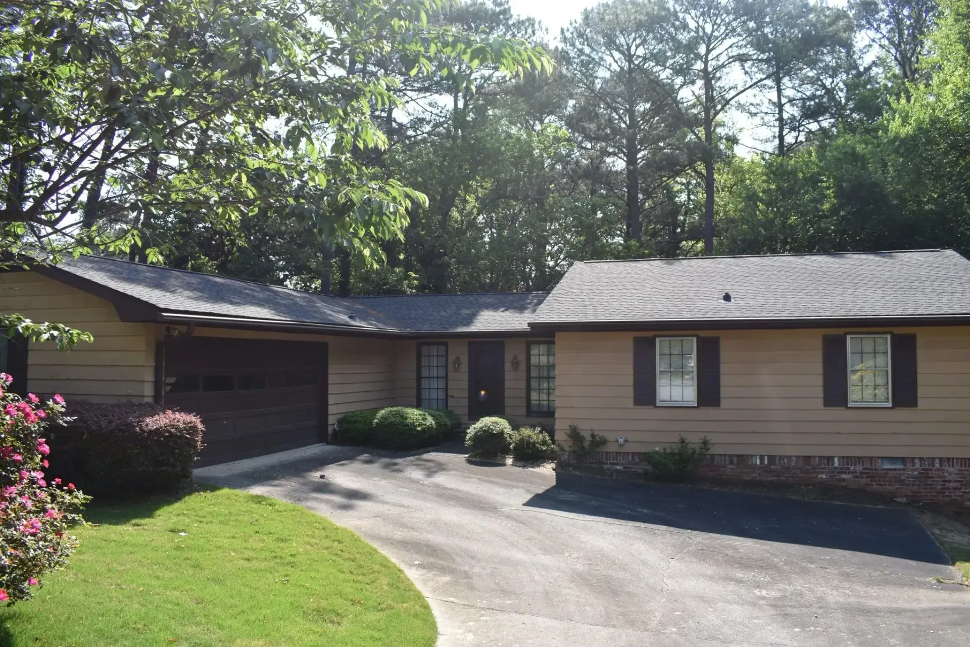 Tan ranch-style house with dark roof and driveway. Trees and bushes surround the property.