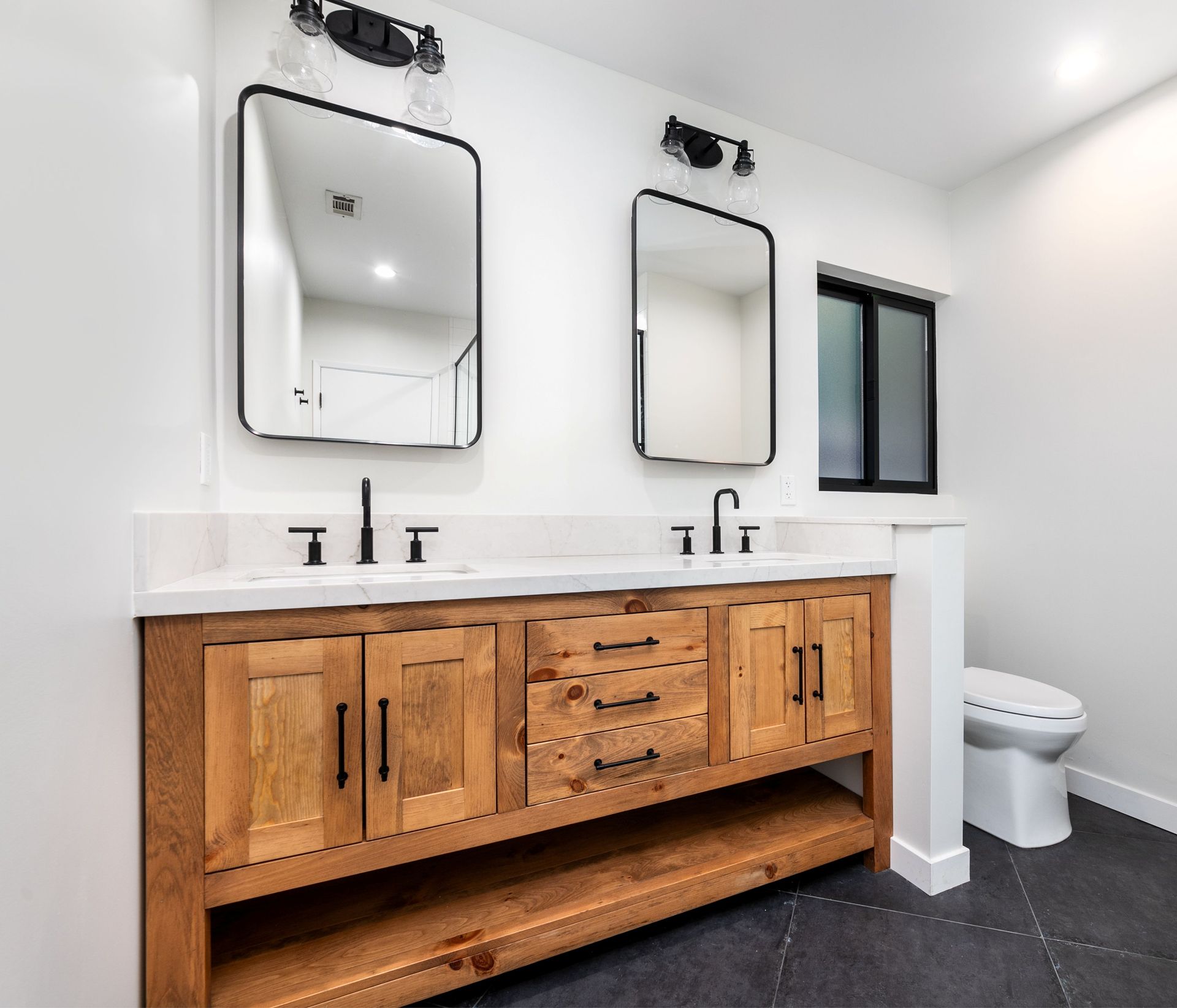 Modern bathroom with double mirrors, wood vanity, black fixtures, and a toilet by a window.
