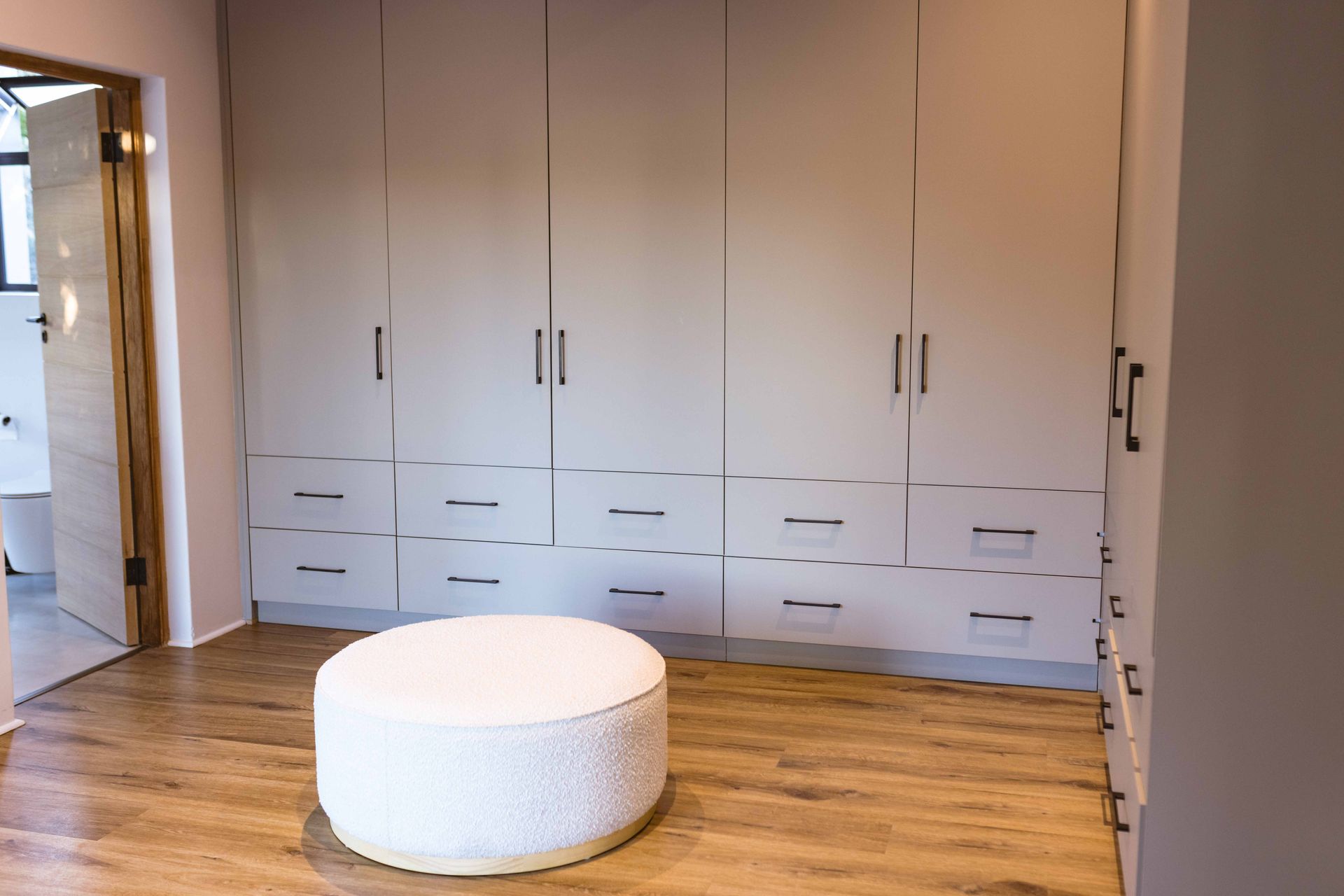 Modern bedroom with built-in beige cabinets, wood floor, and a round white ottoman in the center.