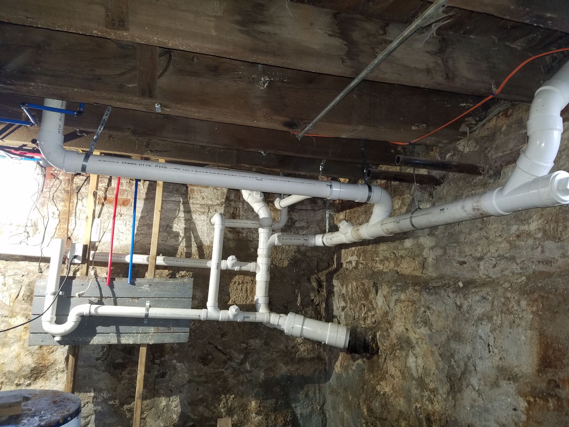 Exposed white plumbing pipes and valves under a wooden floor in a basement.